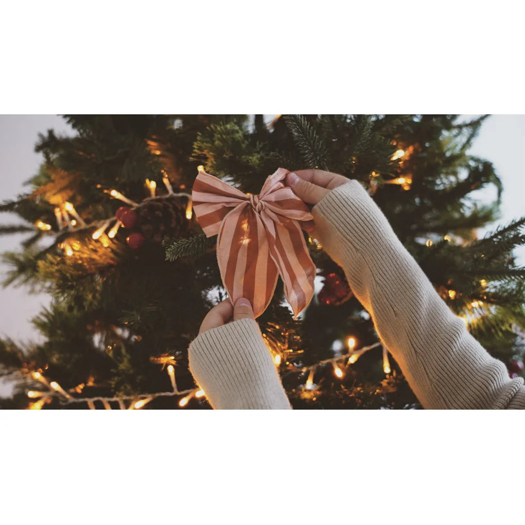 Zwei Hände in kuscheligen Pulloverärmeln befestigen eine rot-weiß gestreifte Schleife von Grech & Co. - Haarmasche an einem Weihnachtsbaum. Der mit Lichtern und Ornamenten von Grech und Co. im Lieblingsmuster-Mix geschmückte Baum sorgt für festliche Stimmung.