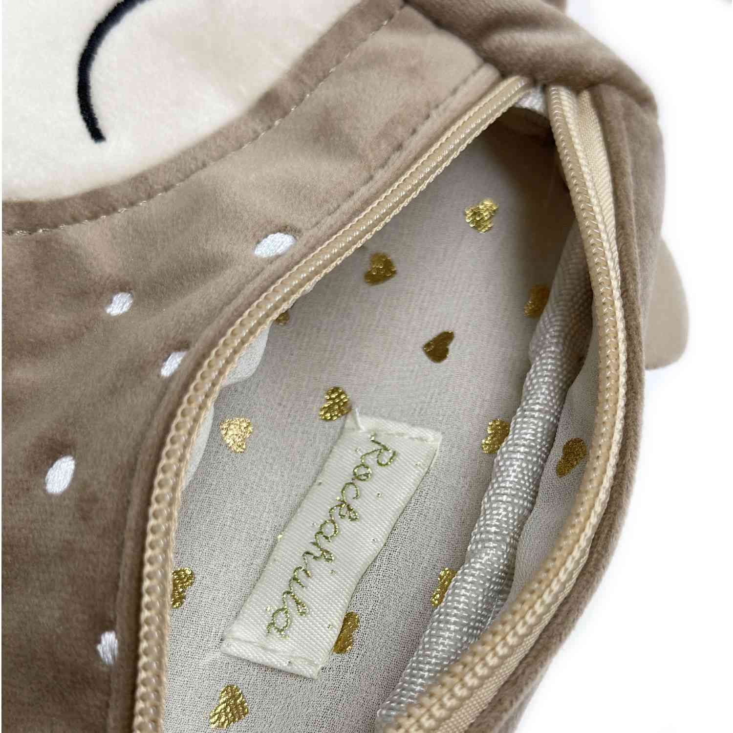 Nahaufnahme eines offenen Reißverschlusses an einem beigen Plüschartikel, der ein weißes Stofffutter mit goldenen Herzmustern im Inneren offenbart. Auf dem Etikett im Inneren steht „Rockahula“, bekannt für entzückende Accessoires, die sich perfekt mit Frisuren bei Kindern kombinieren lassen. Bei diesem besonderen Artikel handelt es sich um die Rockahula - Rehntier Bauchtasche.