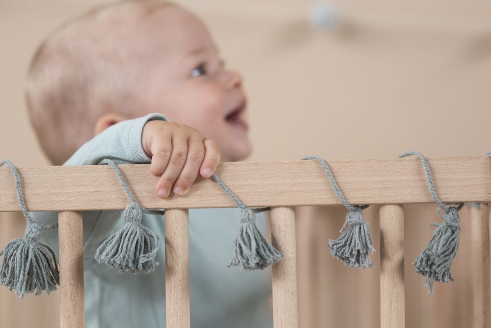 Ein Baby steht in einem Holzbettchen, das mit Schnüren von Lorena Canals geschmückt ist – Girlande Neverending Story von Lorena Canals, was eine bezaubernde Dekoration für das Kinderzimmer schafft. Das lächelnde Baby in einem hellblauen Outfit klammert sich mit einer Hand an das Gitter des Bettchens und schaut zur Seite. Der weiche, neutrale Hintergrund unterstreicht diesen ruhigen Moment.