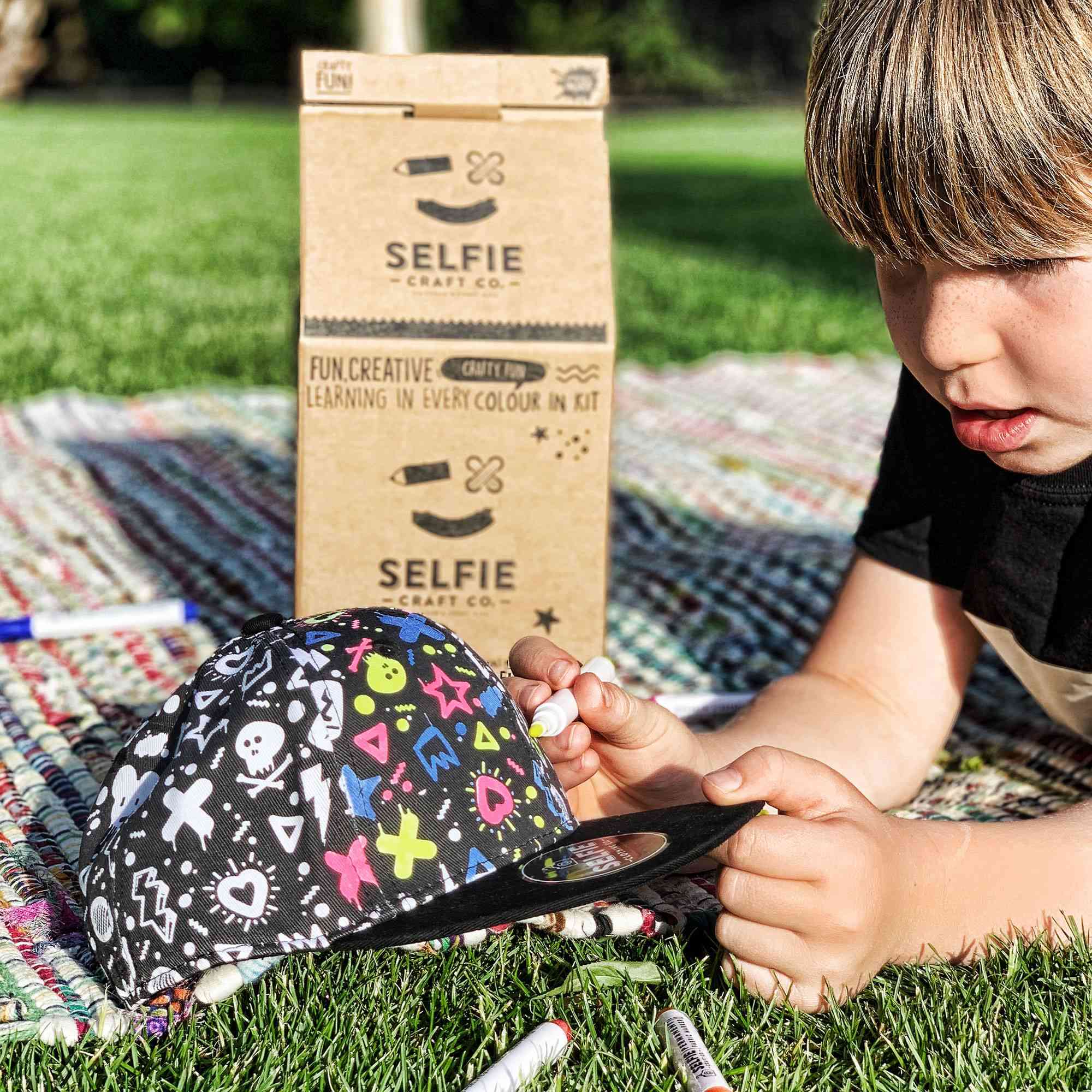 Ein Kind liegt im Gras und bemalt eine schwarze Kappe mit bunten Formen und Motiven von Selfie Craft Co. – Graffiti Colour in Snapback Kappe. Im Hintergrund ist ein Karton mit der Aufschrift „Selfie Craft“ mit Anleitungen und Beschreibungen zu sehen. Die Szene scheint an einem sonnigen Tag im Freien zu spielen, umgeben von Bastelutensilien für einfaches Basteln mit Kindern.