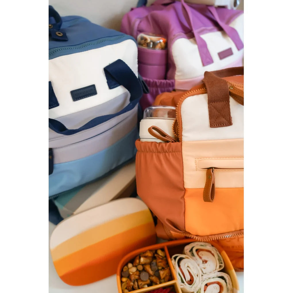 Ein übersichtlich geordnetes, buntes Sortiment mit drei Rucksäcken in Blau, Lila und Orange, jeweils mit Taschen und Reißverschlüssen, einer gestreiften Lunchbox, einer Grech und Co. – Grosse Lunchbox aus Silikon für die Snacks im Kindergarten und einem gelben Tablett mit gerollten Wraps und Leckereien.