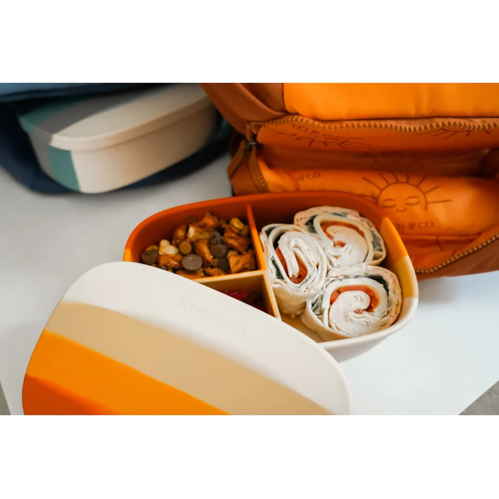 Eine Grech & Co. - Grosse Lunchbox aus Silikon mit einem orange, gelb und weiß gestreiften Deckel. Der Behälter enthält Tortilla-Windräder mit sichtbarer Füllung, kleine Brezeln und eine Mischung aus Schokoladenstückchen und Trockenfrüchten. Im Hintergrund ist neben einer Grech und Co. Lunchbox eine orangefarbene Tüte mit Sonnenmotiv zu sehen.