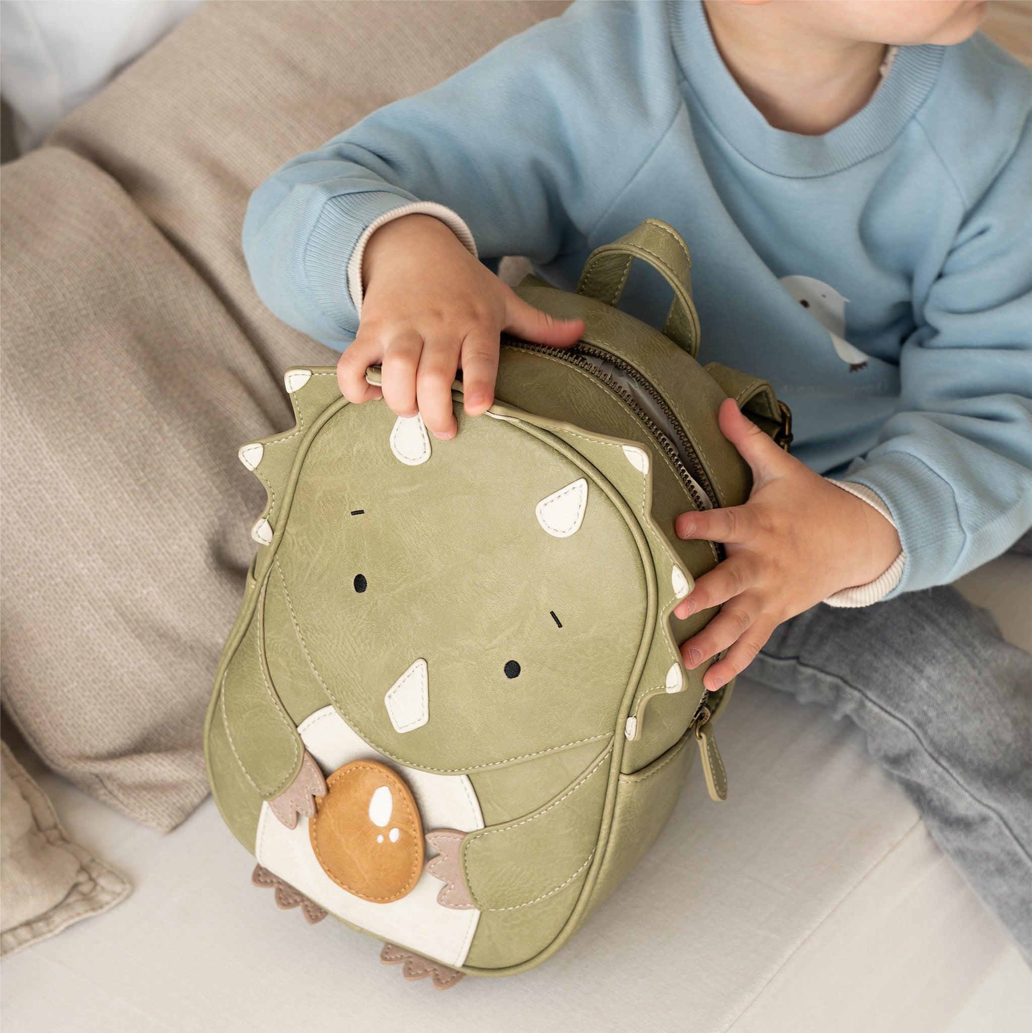 Ein Kind in einem blauen Pullover sitzt auf einem Sofa und hält einen nachhaltig hergestellten, grünen Rucksack in Dinosaurierform. Der Little Who - Rucksack Dinosaurier Oskar hat weiße Stoffstacheln, ein süßes Dinosauriergesicht und reflektierende Aufkleber. Das Kind öffnet den Reißverschluss zwischen beigen Kissen und weißer Bettwäsche von Little Who.