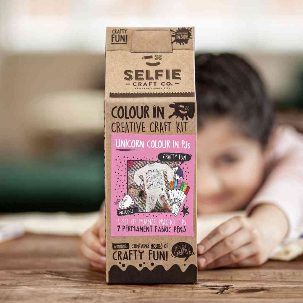 Ein Kind im Hintergrund hält eine Packung Selfie Craft - Selfie Craft Co. - Pyjama in Einhornfarbe, die zum Ausmalen von Einhorn-Pyjamas gedacht ist. Dieses DIY-Set enthält einen Pyjama, 7 permanente Stoffstifte und Übungstipps und verspricht stundenlangen Bastelspaß für Kinder.