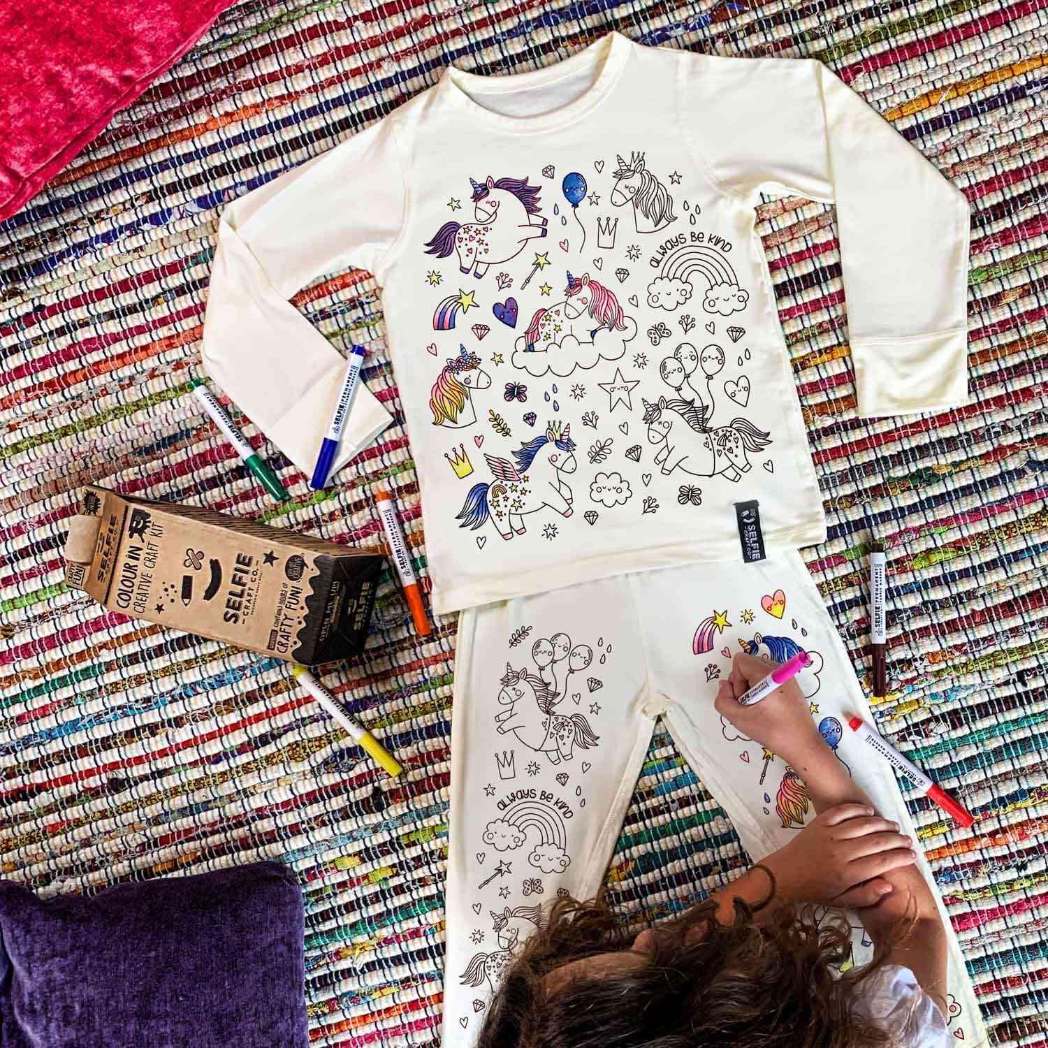 Ein Kind malt Bilder mit Einhornmotiven auf seinem weißen Selfie Craft Co.-Pyjama in Einhornfarbe aus, während es auf einem bunten gewebten Teppich liegt. Der Pyjama zeigt verschiedene umrissene Illustrationen wie Einhörner, Luftballons und Wolken. Farbige Stifte aus einem Selfie Craft-Bastelset liegen verstreut herum, und eine Stiftebox liegt in der Nähe.