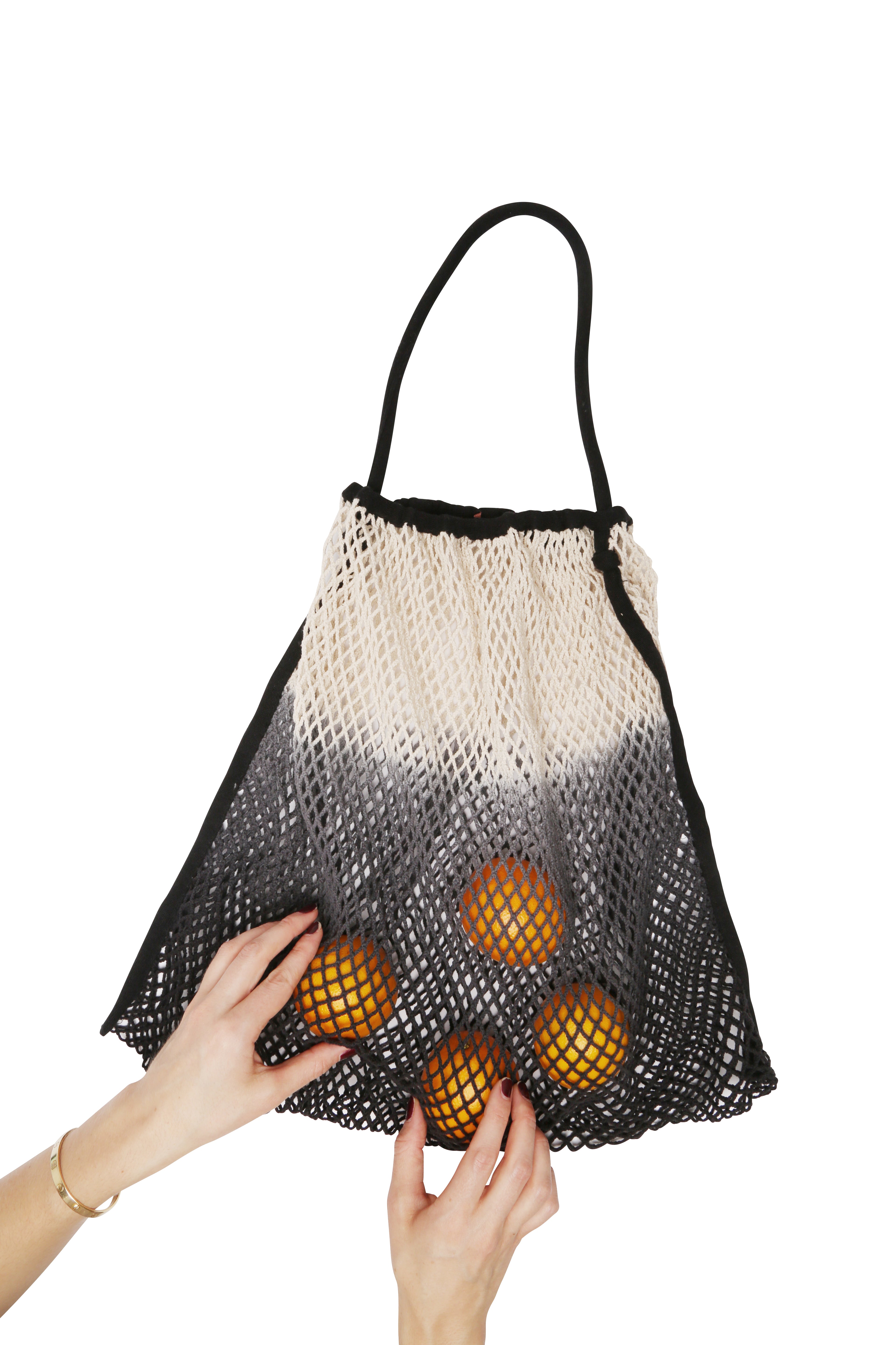 Die Hände einer Person halten einen schwarzen Bären von Mara Mea mara mea - Kinderwagen Netz -, der mehrere Orangen enthält. Die Tasche hat ein Ombre-Design mit Schwarz unten, das nach oben in Weiß übergeht, was eine nachhaltige Wahl für Käufer verkörpert. Der Hintergrund ist schlicht weiß.