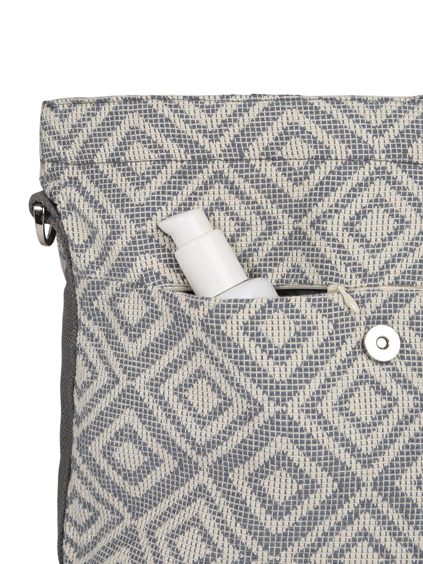 Nahaufnahme eines Stoffbeutels mit grau-weißem geometrischem Muster. In einer Außentasche ist teilweise eine kleine Flasche mit Pumpverschluss zu sehen, die mit spülmaschinenfesten Aufklebern versehen ist. Die Tasche hat einen Metallknopfverschluss beim Mara Mea mara mea - City Wickelrucksack - extra Romantik.