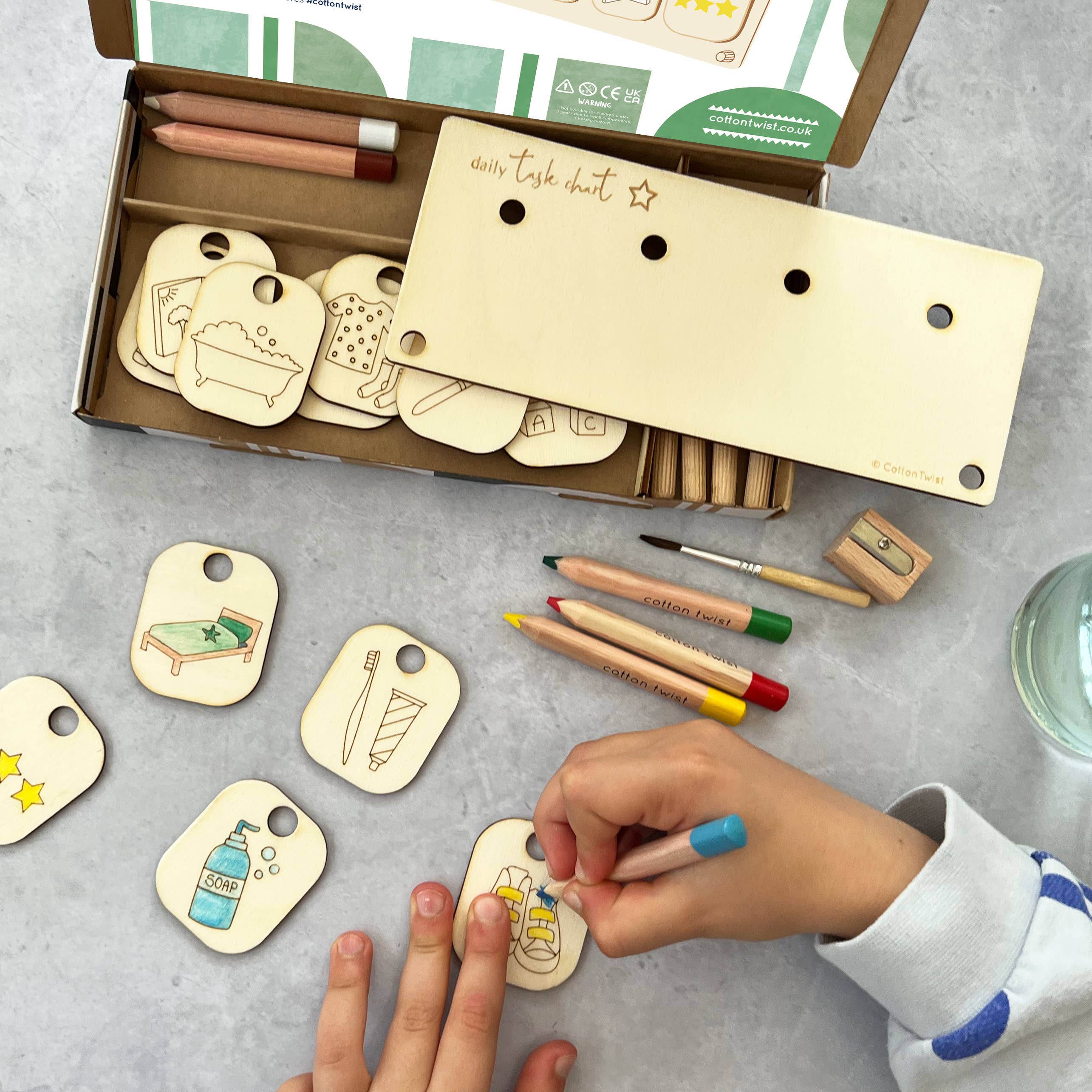 Mit dem Cotton Twist - Aufgabenroutine-Set von Cotton Twist verfeinert ein Kind seine Feinmotorik, indem es an einem Tisch Holzschilder mit Abbildungen mit Buntstiften ausmalt. Um sie herum befinden sich weitere plastikfreie Holzschilder mit Zeichnungen wie einer Flasche und einer Badewanne sowie einer offenen Schachtel und einem Holzbrett mit Löchern – perfekt zum Gestalten umweltfreundlicher Partygeschenke.