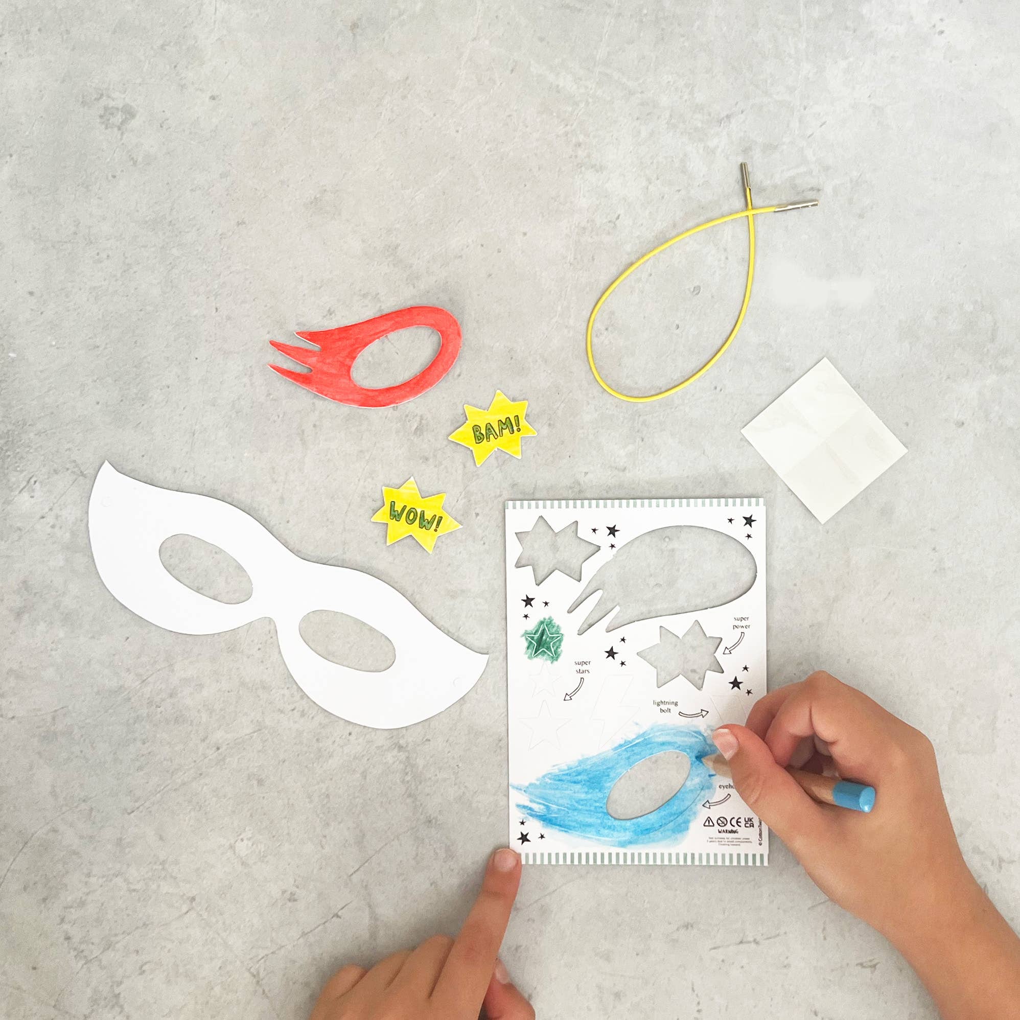 Man sieht die Hände eines Kindes, das eine Zeichnung mit Superheldenmotiv auf Papier ausmalt. Neben der Zeichnung sind verschiedene ausgeschnittene Papierformen zu sehen, darunter eine weiße Cotton Twist - DIY Superhelden-Maske, eine rote Flamme und gelbe Comic-Wörter wie „BAM!“ und „WOW!“. Ein Lineal, Schnur und Haftnotizen aus umweltfreundlichen Materialien sind ebenfalls zu sehen.