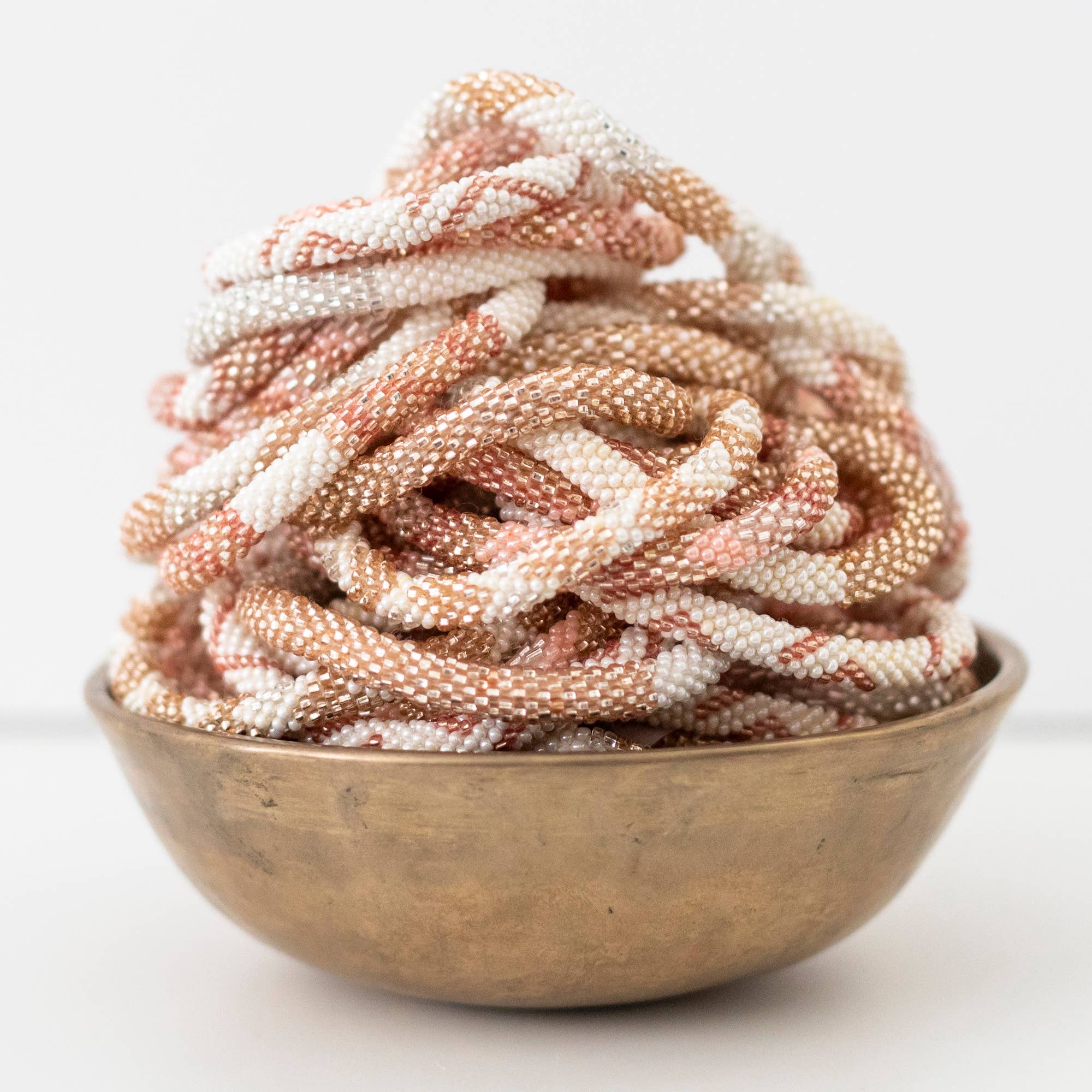 Ein aufgerollter Stapel aufwendig mit Perlen besetzter Aid Through Trade - Armbänder Damen in Gold-, Weiß- und Roségoldtönen sitzt elegant in einer rustikalen Messingschale. Die kleinen, gleichmäßigen Perlen sorgen für ein strukturiertes, luxuriöses Erscheinungsbild. Diese handgefertigten Armbänder von Aid Through Trade werden vor einem sauberen, schlichten weißen Hintergrund präsentiert.