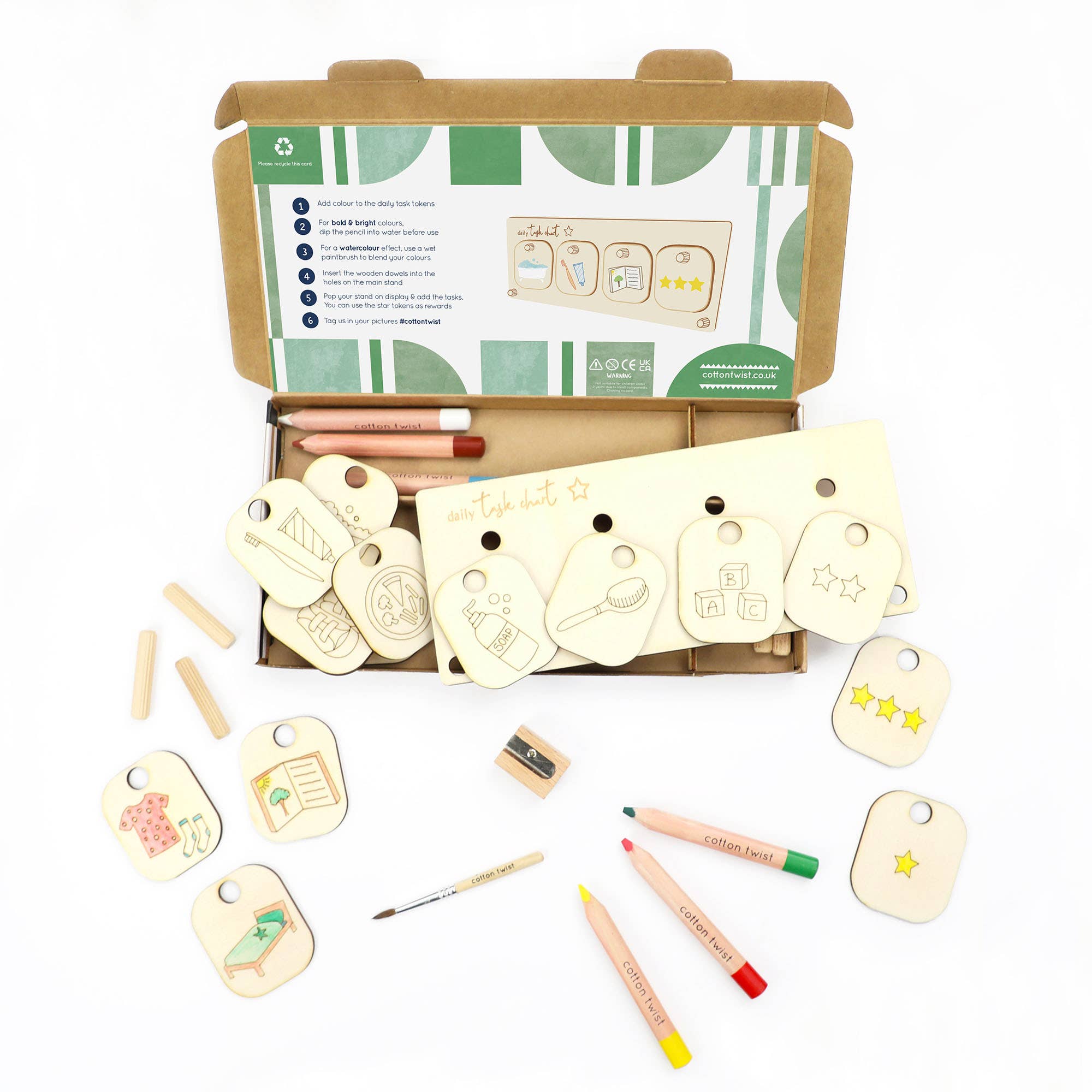 Die Cotton Twist - Aufgabenroutine von Cotton Twist wird auf einer weißen Oberfläche präsentiert und zeigt eine offene Box mit Anweisungen auf der Unterseite des Deckels. Dieses plastikfreie Set enthält Holzplättchen mit verschiedenen Abbildungen, Holzstifte, Buntstifte und einen Anspitzer. Die Gegenstände sind ordentlich um die Box herum angeordnet.