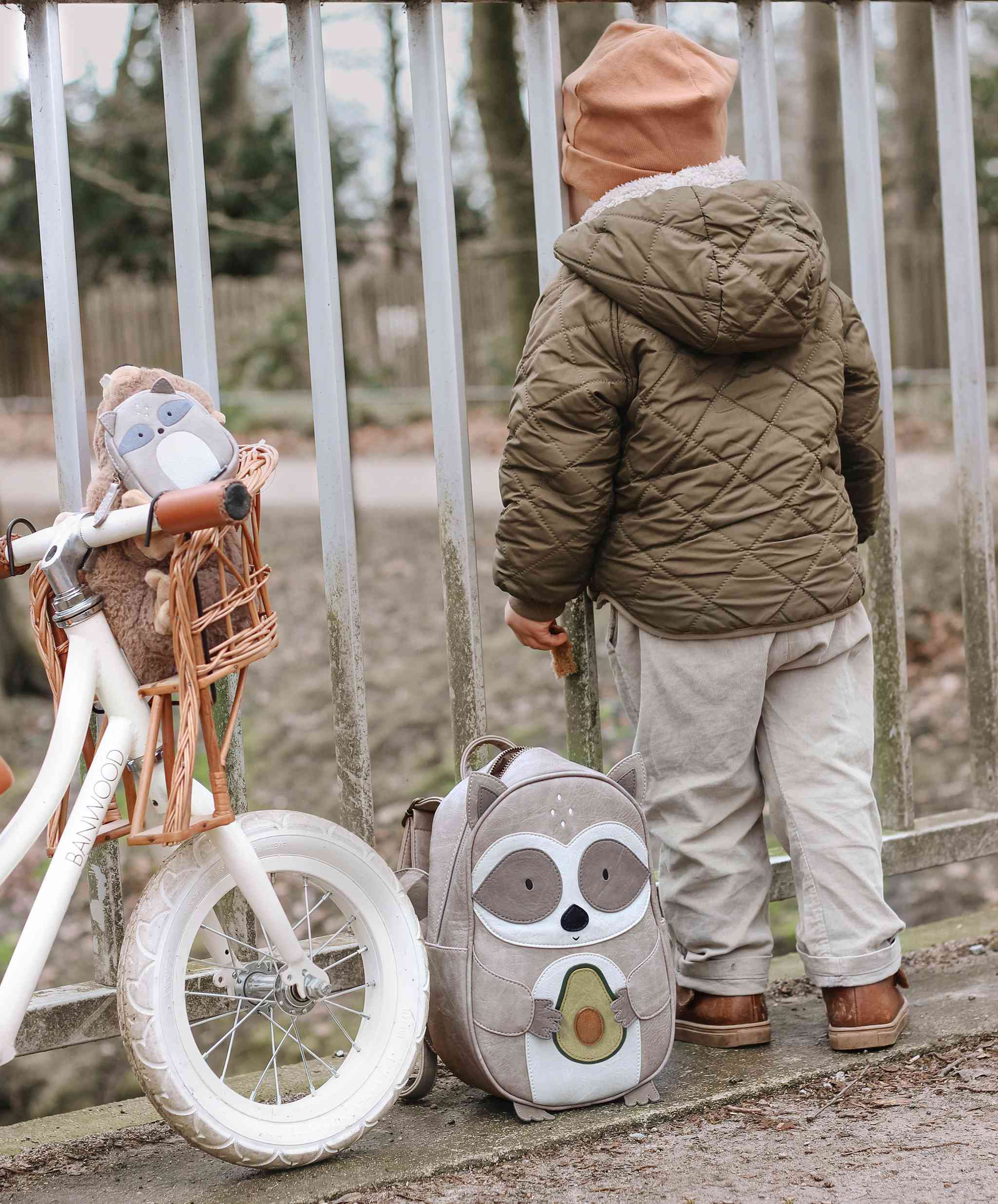 Ein Kind in einer braunen Steppjacke und hellbraunen Hosen schaut durch einen Zaun. Neben dem Kind steht ein weißes Laufrad mit einem Plüschtier im Korb, geschmückt mit spülmaschinenfesten Aufklebern. Auf dem Boden liegt ein Little Who - Rucksack Waschbär Waldemar von Little Who, mit einem Cartoon-Eulen-Design. Die Szene spielt im Freien mit Bäumen im Hintergrund.