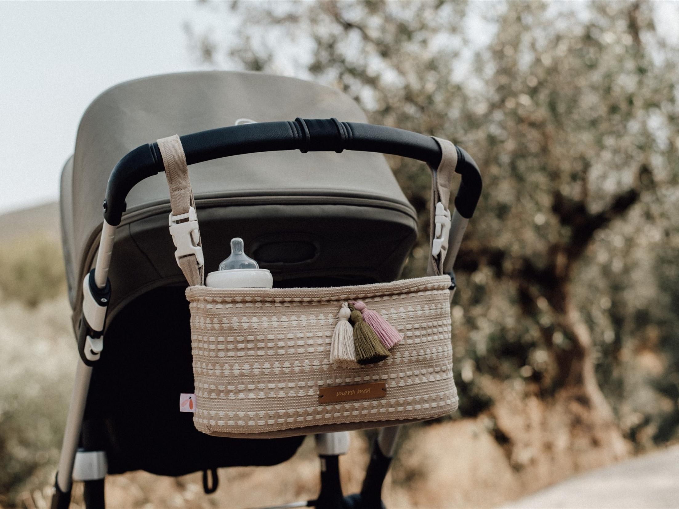 Ein Kinderwagen mit einem hellbraunen und weißen geflochtenen Organizer, der am Griff hängt. Der Mara Mea Mara Mea - Kinderwagen-Organizer - Seashore Grove, perfekt für jedes Kinderartikelgeschäft, bietet Platz für eine Babyflasche und ist mit bunten Quasten geschmückt. Der Hintergrund zeigt eine Außenkulisse mit verschwommenen Bäumen und Blättern.