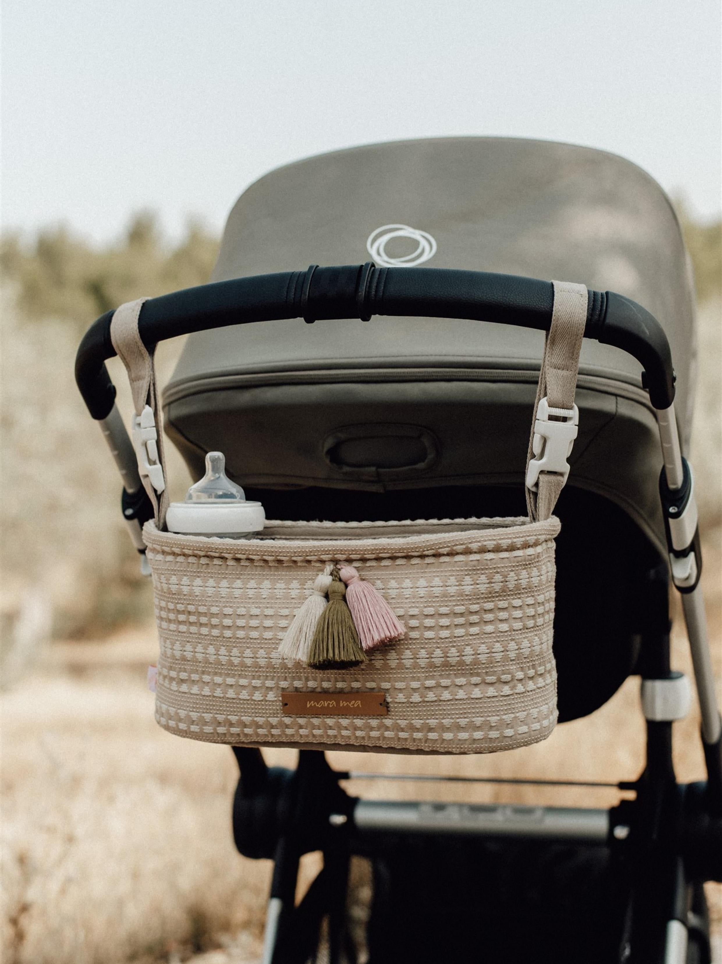Ein Kinderwagen mit einem in neutralen Farben gehaltenen Aufbewahrungsorganizer aus Strick, der an der Lenkstange hängt, ist im Freien abgebildet. Der Mara Mea - Kinderwagen-Organizer - Seashore Grove von Mara Mea hält eine Babyflasche und verfügt über Quasten in Beige, Grün und Rosa, die wahrscheinlich in Ihrem örtlichen Kinderartikelgeschäft erhältlich sind. Der Hintergrund zeigt eine verschwommene Naturkulisse.