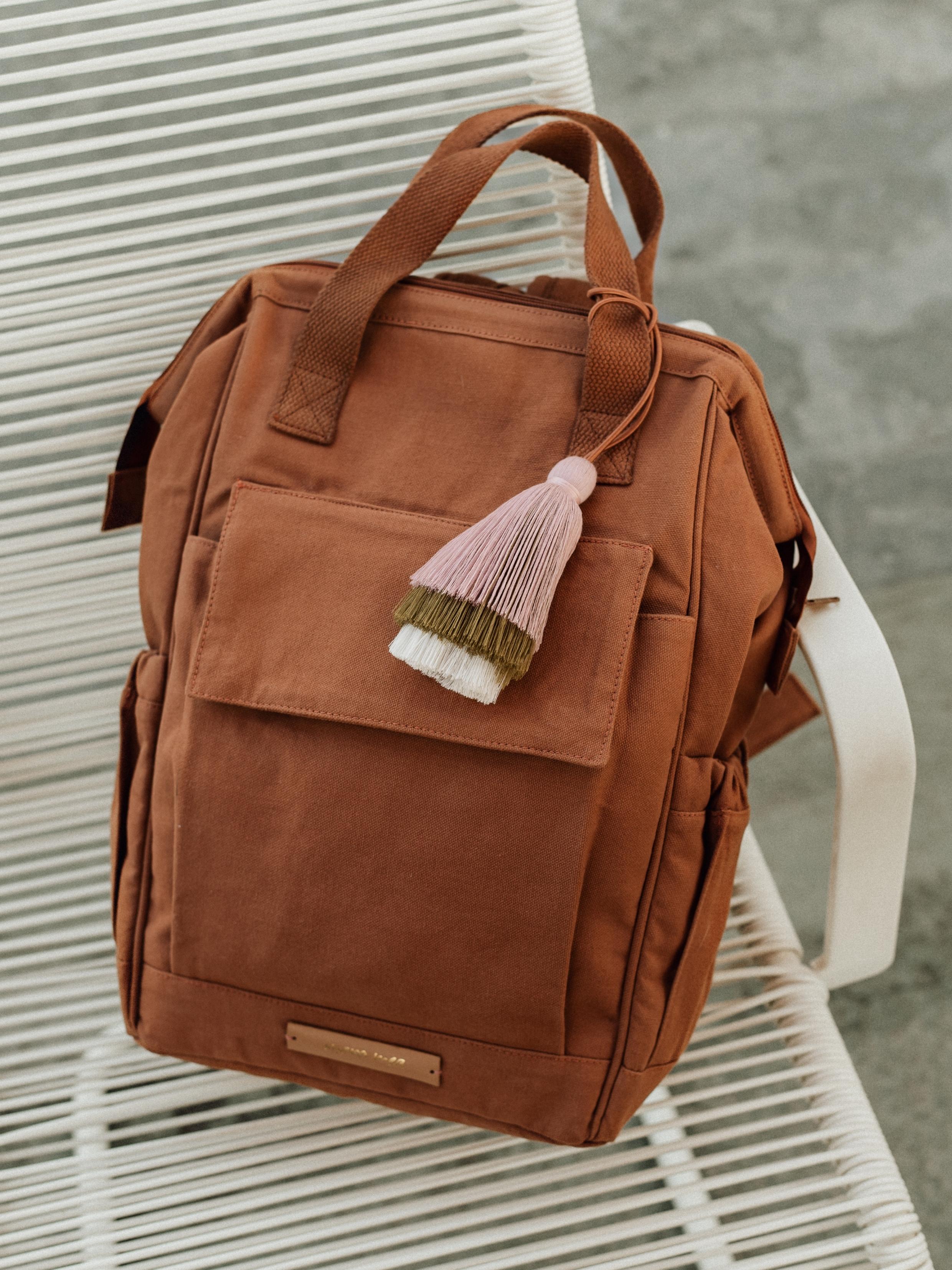 Ein brauner Mara Mea - Travel Wickelrucksack - Desert Fox steht auf einem weißen Lattenstuhl. Der im kinderartikelgeschäft erhältliche Rucksack hat mehrere Taschen und einen Tragegriff an der Oberseite. Am Reißverschluss ist eine dekorative Quaste in Rosa-, Weiß- und Grüntönen befestigt. Der Hintergrund ist eine neutrale Betonoberfläche.