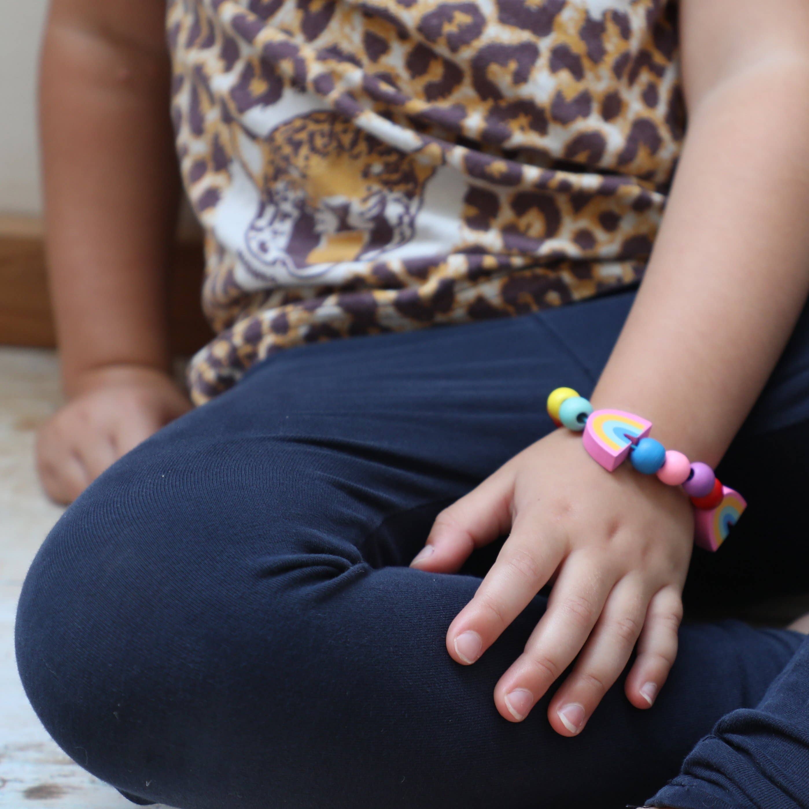 Ein Kind sitzt mit übereinandergeschlagenen Beinen auf dem Boden und trägt dunkelblaue Leggings und ein Shirt mit Leopardenmuster. An seinem Handgelenk trägt es ein buntes, plastikfrei Cotton Twist – DIY Regenbogen-Armband, das mit Herzformen verziert ist. Die Hand des Kindes ruht sanft auf seinem Knie.