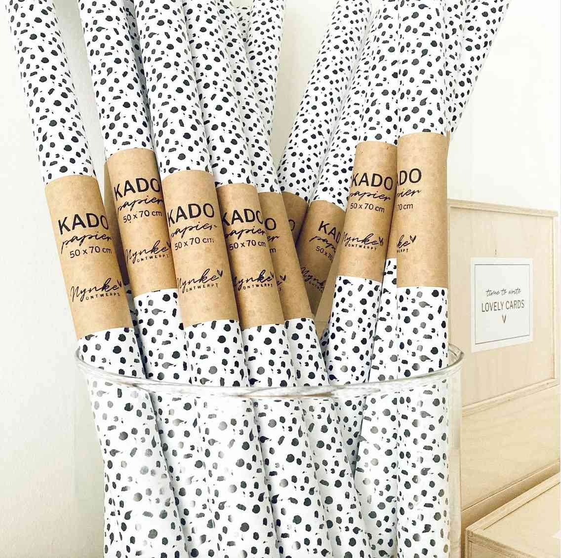 Ein durchsichtiger Behälter ist mit Rollen Nynke Ontwerpt - Geschenkpapier Punkte gefüllt, jede mit der Aufschrift „50x70 cm“. Das weiße Papier weist ein schwarz gesprenkeltes Punktmuster auf. Im Hintergrund befindet sich eine Holzschachtel mit einem Etikett mit der Aufschrift „LOVELY CARDS“, perfekt für eine bezaubernde Geburtstagskarte.