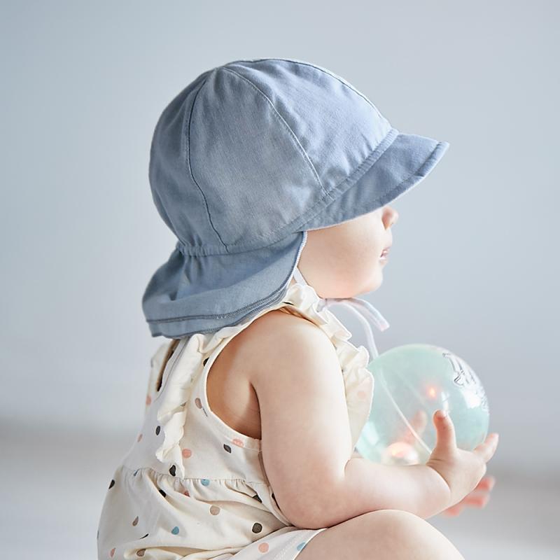 Ein Baby in einem hellen Outfit mit Punkten und einer blauen Maximo - BABY-Schildmütze von Maximo hält einen durchsichtigen Ball. Das Baby blickt abgewandt, sanftes, natürliches Licht erhellt die Szene vor einem einfachen Hintergrund – und fängt so perfekt die Essenz der Frühlings-/Sommersaison ein.