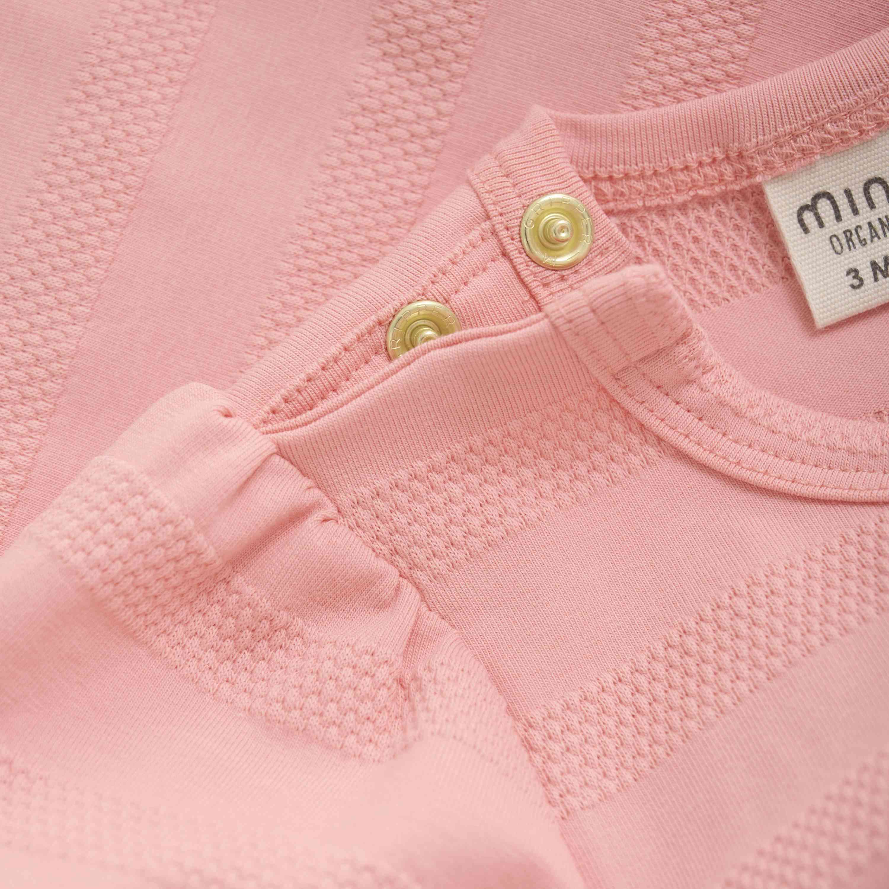 Nahaufnahme eines rosa Babybodys mit strukturierten horizontalen Streifen. Der Ausschnitt ist mit zwei goldenen Druckknöpfen versehen, die das Anziehen erleichtern. Ein weißes Etikett im Inneren des Kleidungsstücks zeigt den Markennamen Minymo und Größeninformationen. Der Bambusstoff wirkt weich und bequem.