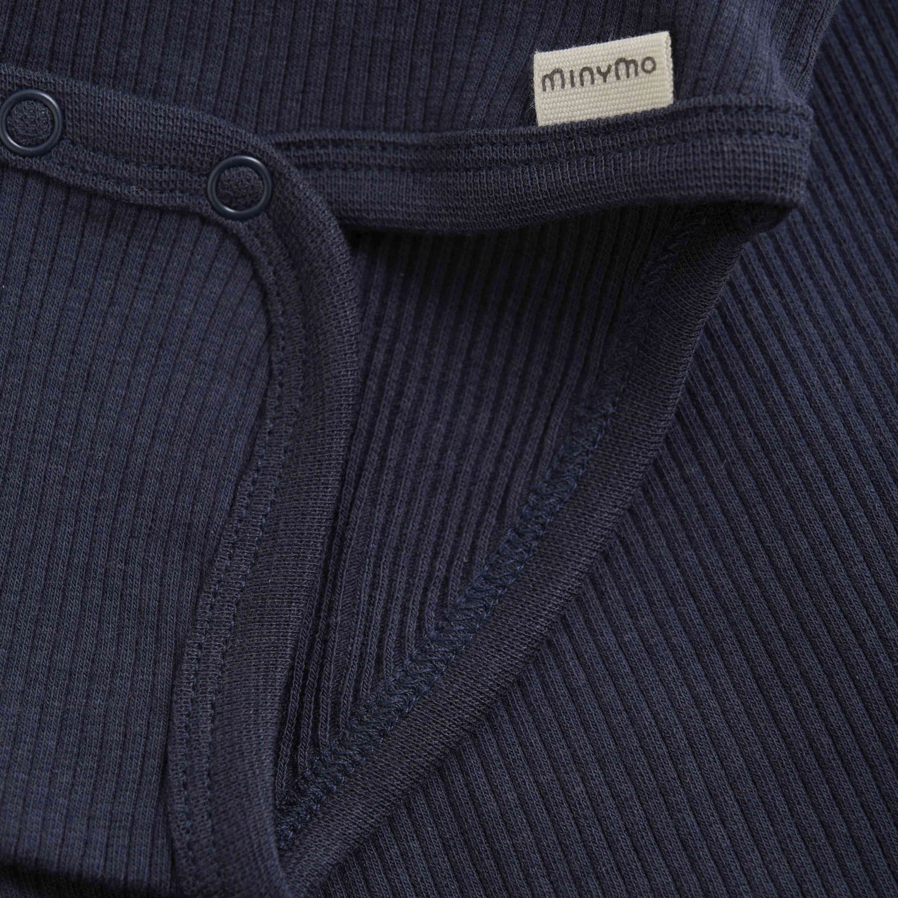 Nahaufnahme eines marineblauen Rippstoffs Minymo - Body kurzarm aus Rippstoff mit Druckknopfdetail. Ein kleines beiges Etikett mit dem Markennamen „Minymo“ ist in eine der Kanten eingenäht. Die Textur des Stoffes und die Nähte sind deutlich sichtbar. Hergestellt aus weichem, nachhaltigem, GOTS-zertifiziertem Bambus für den Komfort Ihres Babys.
