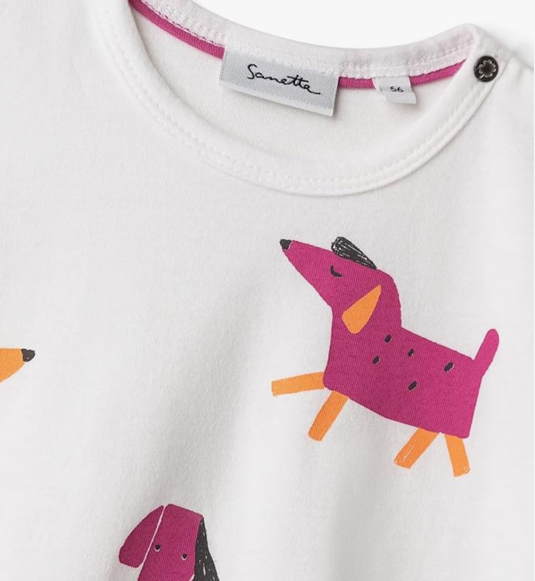 Nahaufnahme eines weißen Sanetta T-Shirts mit verspielten, abstrakten Hundeillustrationen in leuchtenden Rosa-, Orange- und Schwarztönen. Am Ausschnitt ist das Markenlabel „Sanetta“ neben einem Größenetikett mit der Angabe der Größe 56 zu sehen. Der Kragen ist aus weicher Baumwolle gefertigt und verfügt an einer Seite über einen Druckknopf.