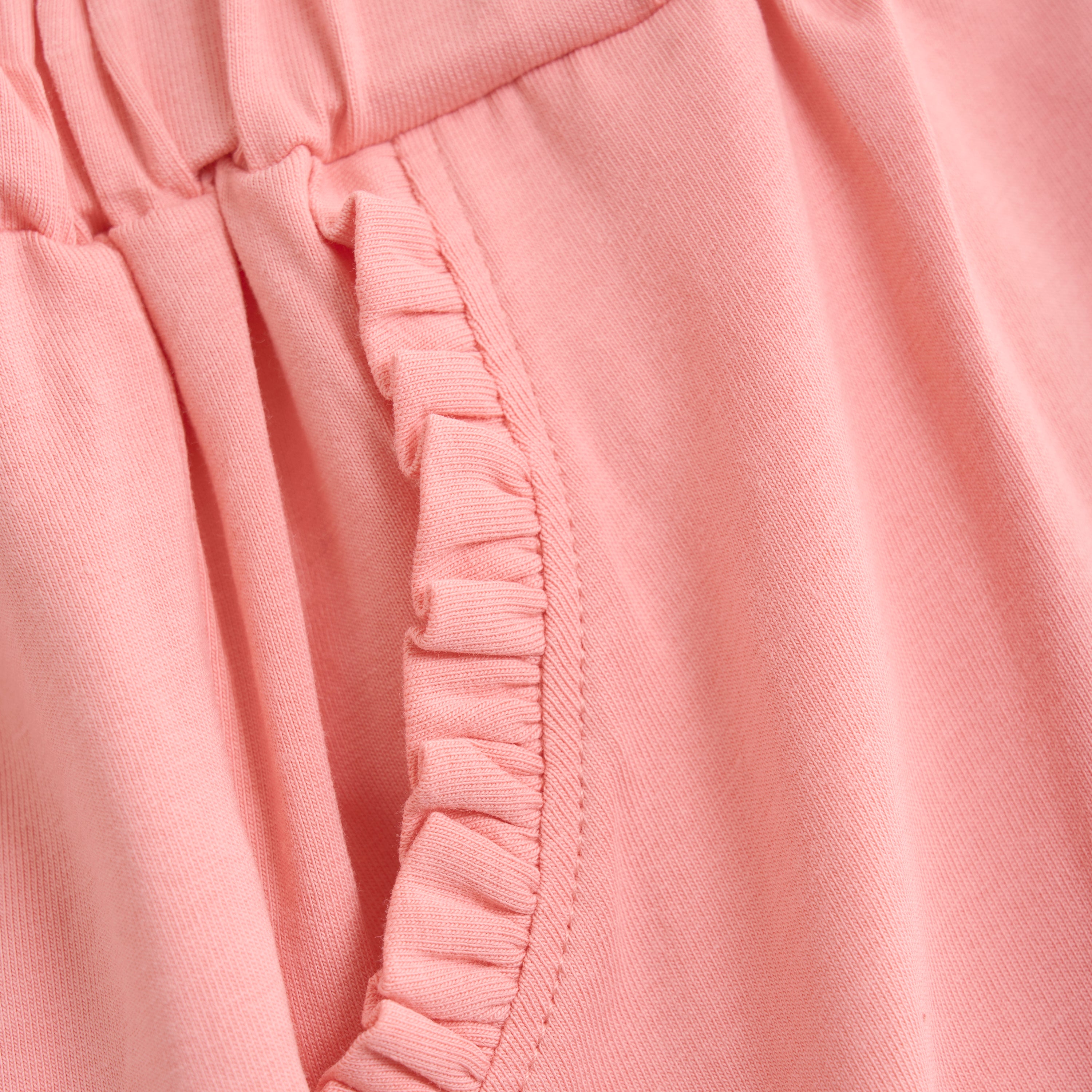 Nahaufnahme der Minymo - Kurze Hosen, die einen rosa Stoff mit gerafftem, elastischem Bund und Rüschenbesatz entlang der Taschenkante zeigt. Die weiche Textur lässt darauf schließen, dass diese Shorts aus bequemem, dehnbarem Baumwollmischgewebe gefertigt sind.