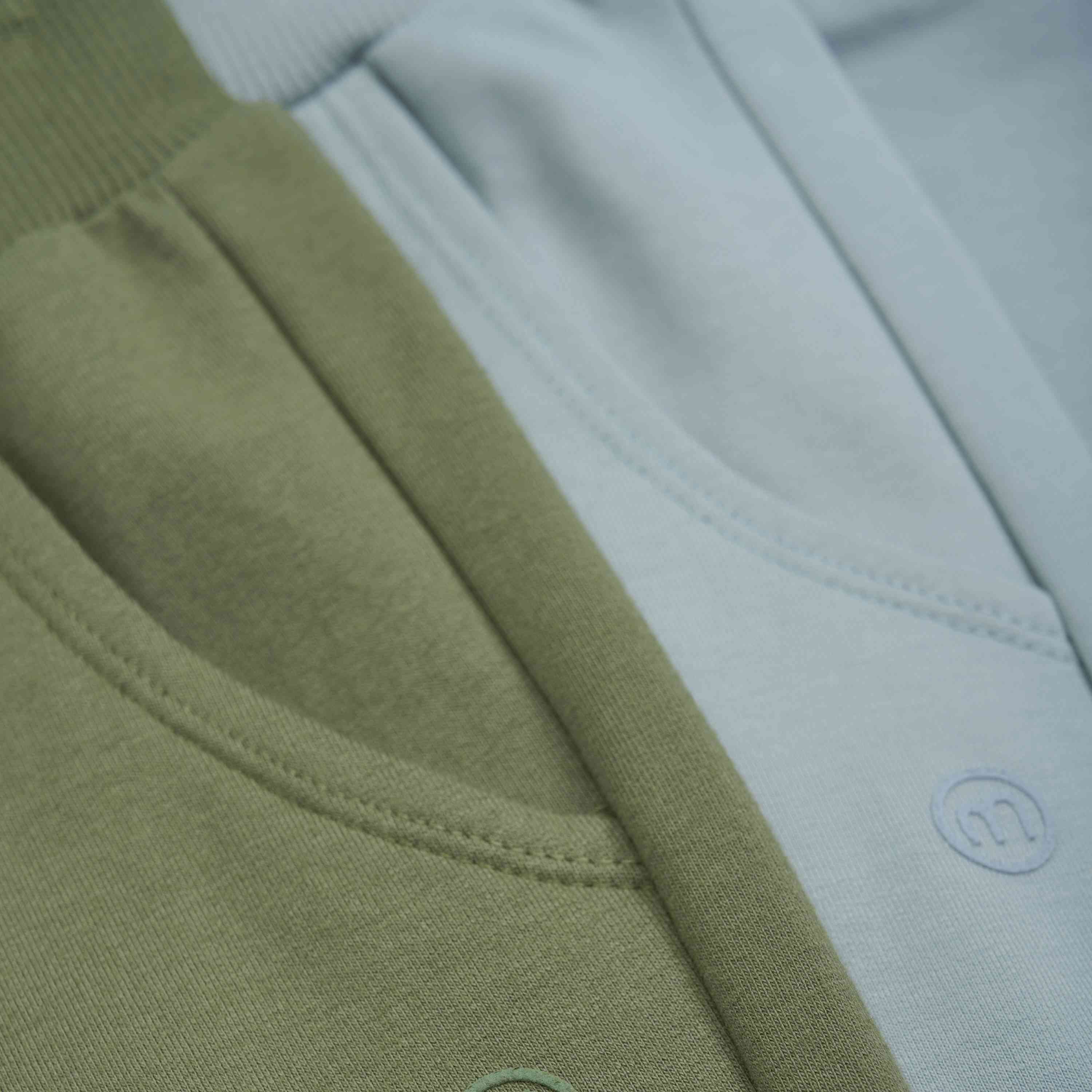 Nahaufnahme detaillierter Teile von zwei Jogginghosen. Eine Hose ist Salbeigrün, die andere Hellblau. Beide haben sichtbare Taschen und gerippte Taillenbänder, die an hochwertige GOTS-zertifizierte Stoffe und Nähte erinnern, die bei Minymo üblich sind – Hosen von Minymo.