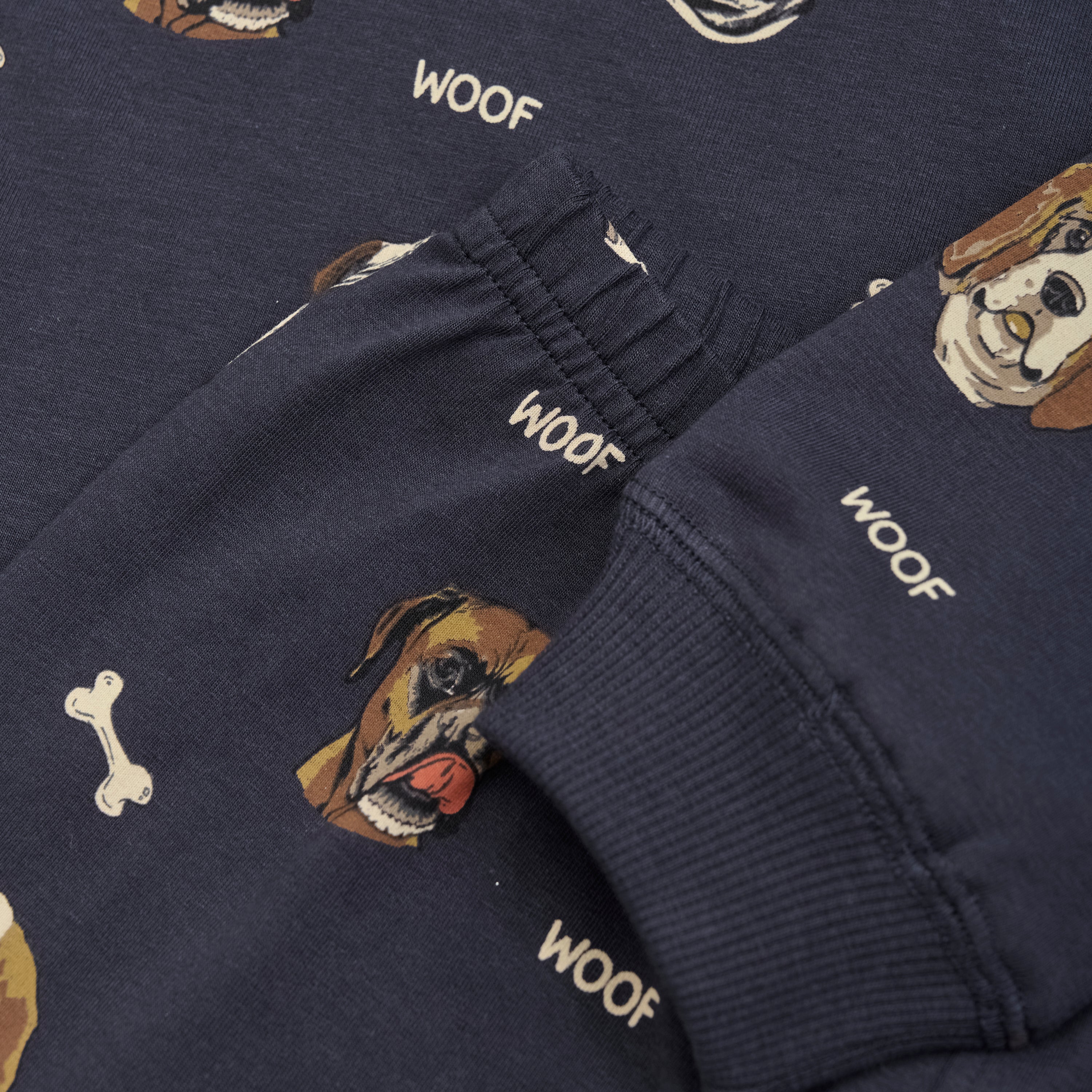 Nahaufnahme eines marineblauen Stoffes mit einem Hundeprint-Nachtwäsche-Muster mit verschiedenen Hundegesichtern, Knochen und dem Wort „WOOF“, das wiederholt in Weiß gedruckt ist. Der Stoff scheint Teil des Minymo-Pyjamas der Marke Minymo zu sein, wahrscheinlich aus einer weichen Baumwollmischung, mit einem sichtbaren Bündchen am Rand.