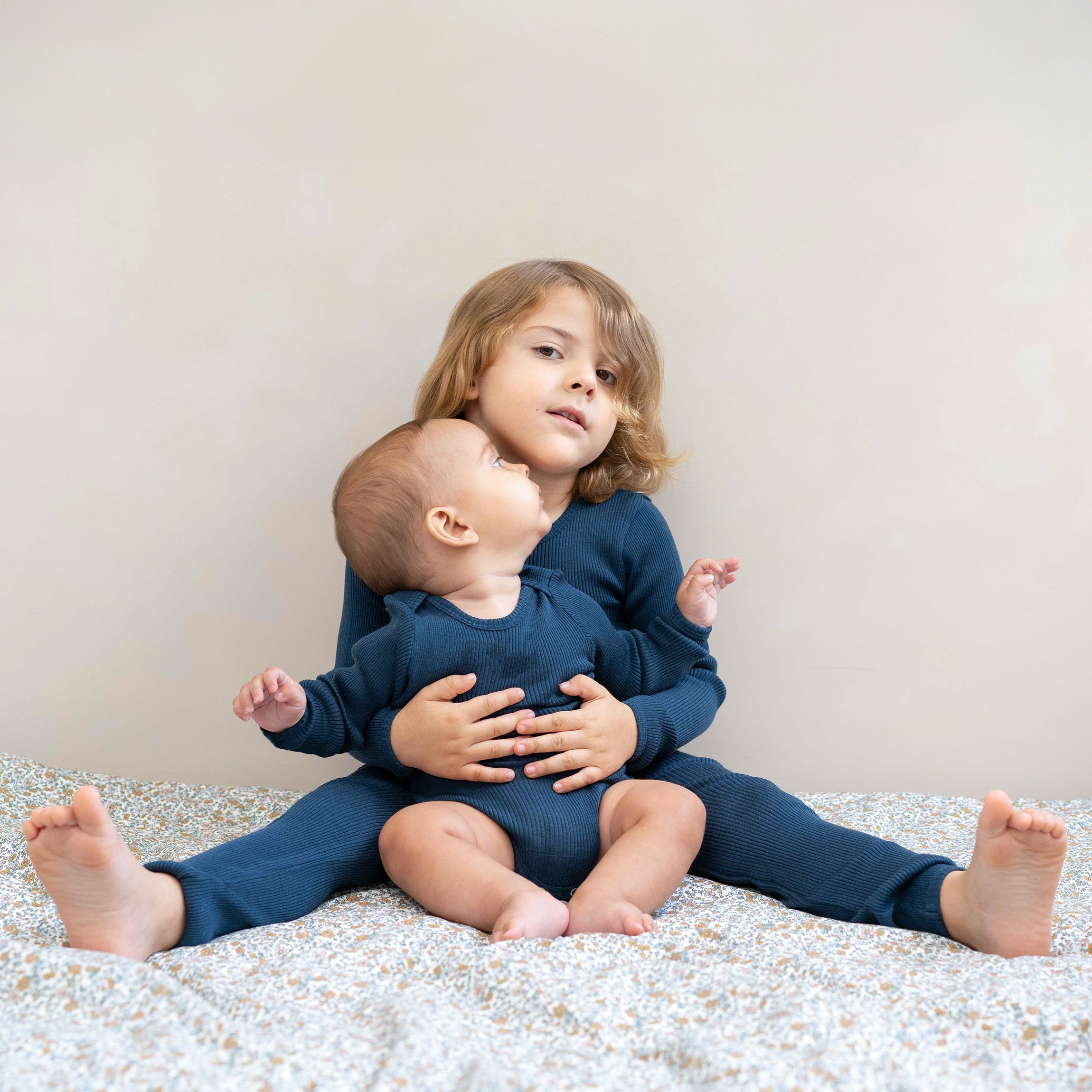 Ein kleines Kind mit langen, hellbraunen Haaren sitzt auf einem Bett und hält ein Baby auf dem Schoß. Beide Kinder tragen passende blaue *minimalisma - Langarmshirts*, die für kuscheligen Wärmekomfort sorgen. Das Baby schaut zu dem älteren Kind auf, während das ältere Kind direkt in die Kamera blickt. Der Hintergrund ist neutral.