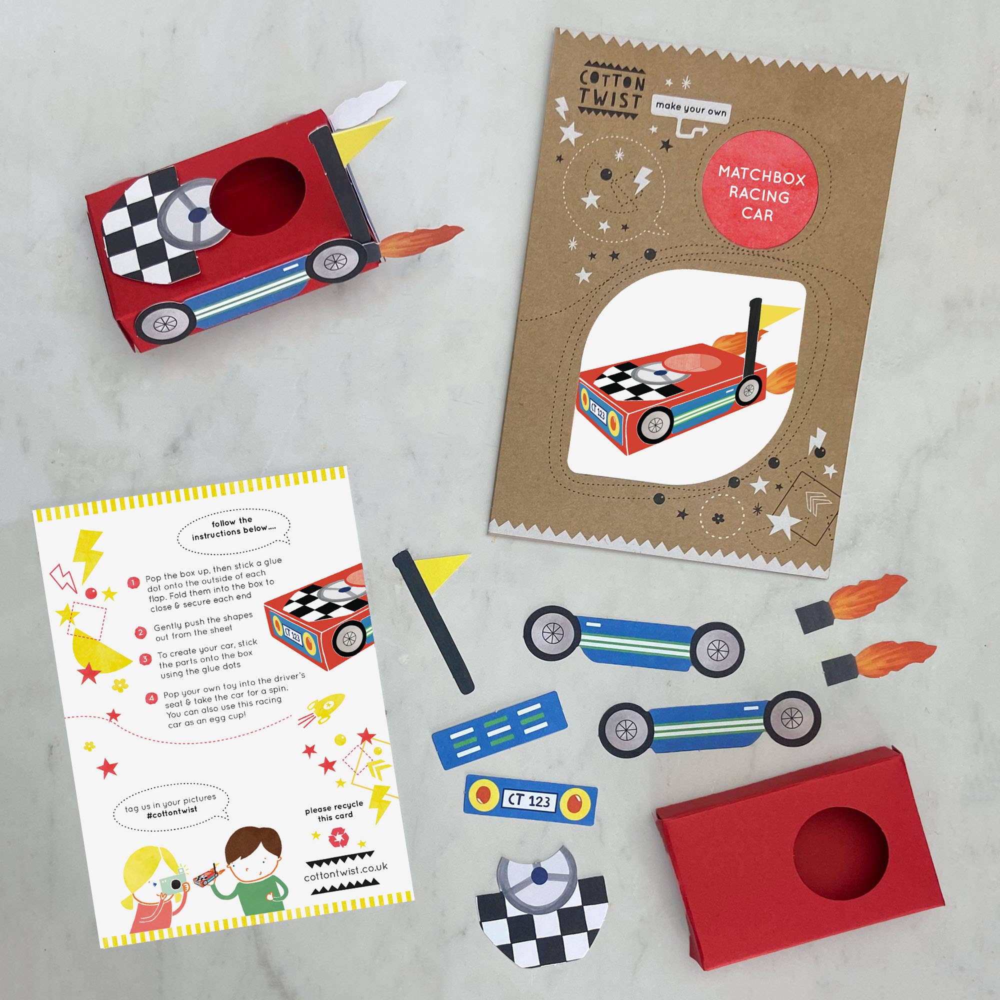 Eine flache Ansicht des Cotton Twist - DIY Matchbox-Rennwagen-Bastelsets zeigt seine Bestandteile: eine rote Schachtel, Aufkleber, Räder, eine Streichholzschachtelhülle und eine bebilderte Anleitung. Die Verpackung ist mit einem Cartoon-Rennwagen und dekorativen Elementen wie Sternen und Flammen verziert. Es eignet sich ideal als Partygeschenk und fördert die Entwicklung der Feinmotorik.