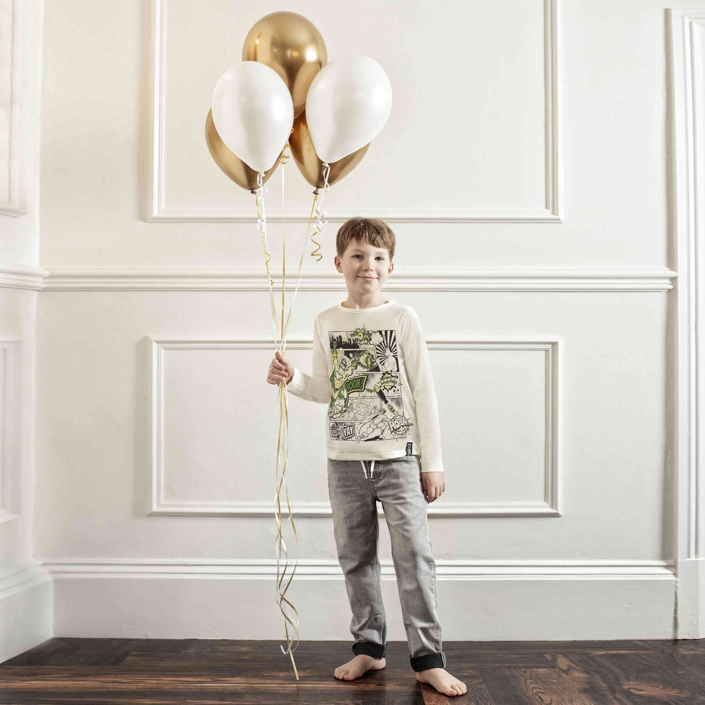 Ein kleiner Junge mit braunem Haar steht drinnen und hält einen Strauß aus drei goldenen und zwei weißen Luftballons an Schnüren. Er ist barfuß und trägt ein weißes Langarmshirt mit einem grün-schwarzen Muster und graue Hosen. Er steht vor einer weiß getäfelten Wand auf einem Holzboden und ist bereit für ein bisschen Selfie Craft – Selfie Craft Co. – Superhelden Comic: Oberteil zum Ausmalen.