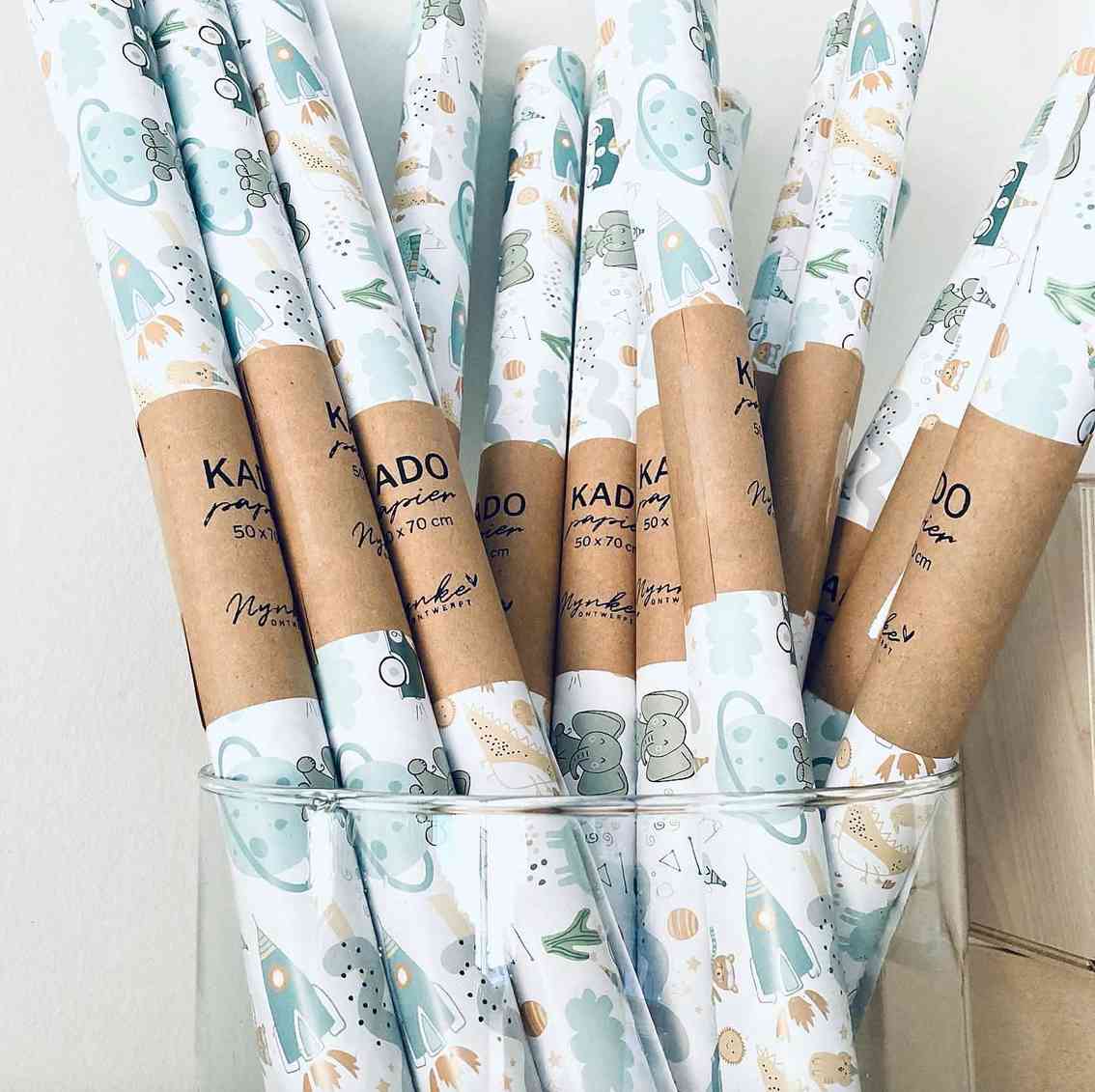 Eine Präsentation von gerolltem Geschenkpapier in einem Halter. Das Papier, das sich perfekt zum Verpacken von Babygeschenken eignet, weist verspielte Designs in Pastellfarben auf, darunter Muster verschiedener Objekte und Formen. Jede Rolle trägt die Aufschrift „Nynke Ontwerpt – Geschenkpapier Kinder Elefant“ mit zusätzlichem Text, der die Abmessungen und andere Details von Nynke Ontwerpt angibt.