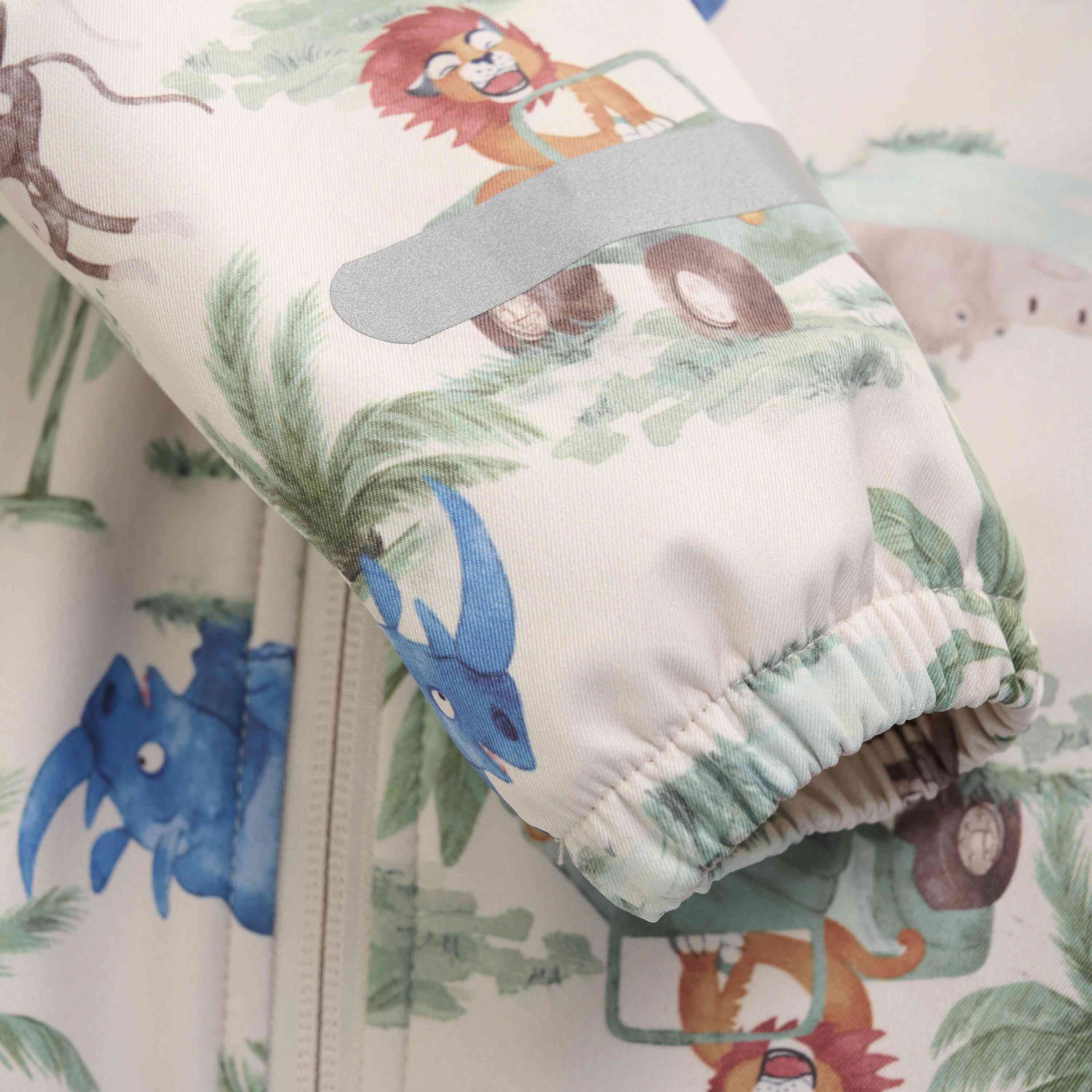 Nahaufnahme eines verspielten Animalprints auf einem CeLaVi - Softshell-Overall von CeLaVi. Das Design umfasst Cartoon-Löwen, die Autos fahren, einen blauen Triceratops und einen Elefanten, alles vor grünem Blattwerk. Der Ärmel des Kleidungsstücks ist sichtbar und zeigt geraffte Manschettendetails – perfekt für jede Waldspielgruppe-Kleiderkollektion.