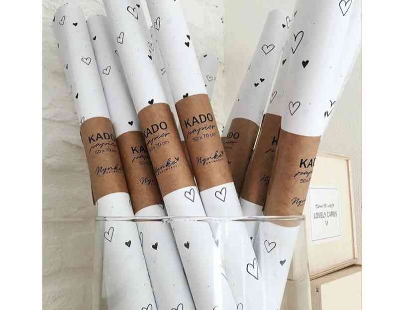 Ein Glasbehälter enthält mehrere Rollen weißes Geschenkpapier mit kleinen schwarzen Herzmotiven. Jede Rolle ist mit einem braunen Etikett aus Kraftpapier umwickelt, auf dem „Nynke Ontwerpt – Geschenkpapier Herzen“ steht und die Größenangaben enthält. Im Hintergrund steht auf einem Schild „Lovely Cards“, ideal zum Kombinieren mit Geburtskarten oder Babygeschenken von Nynke Ontwerpt.
