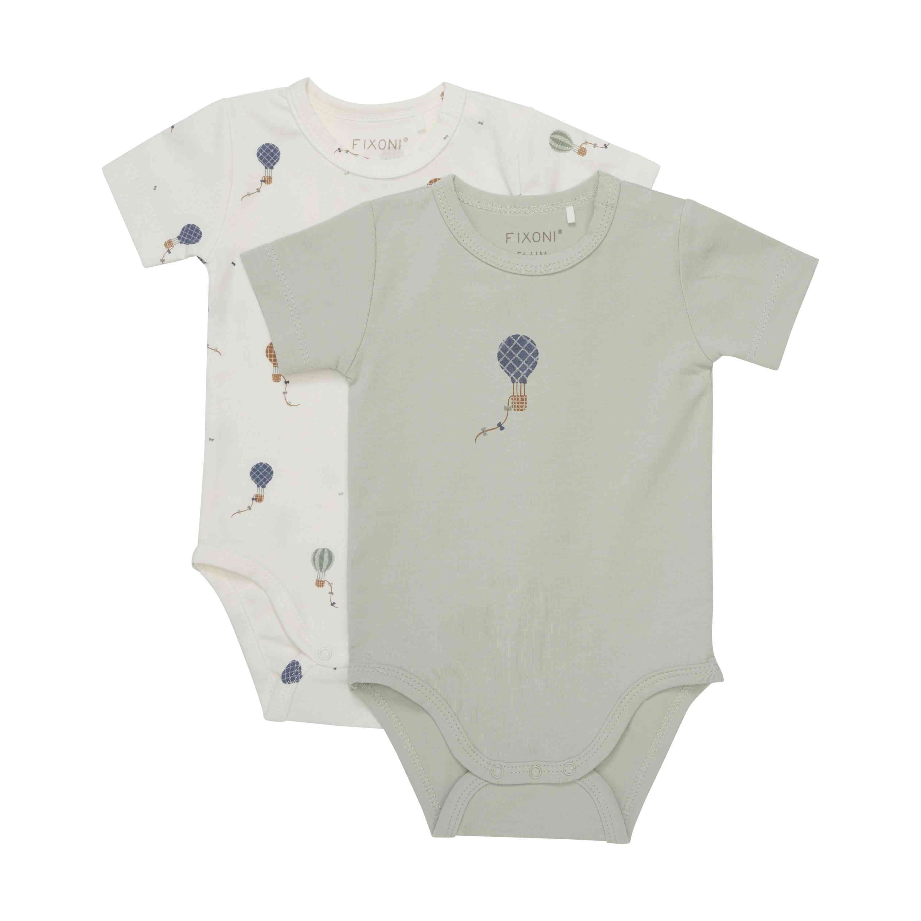 Auf weißem Hintergrund sind zwei Babybodys abgebildet. Der vordere Babybody ist salbeigrün mit einem Heißluftballon-Design. Der hintere Babybody ist weiß mit einem Muster aus kleinen Heißluftballon-Illustrationen. Beide Kleidungsstücke sind kurzärmlig und im Fixoni-Stil gefertigt, perfekt als Fixoni - Body kurzarm.