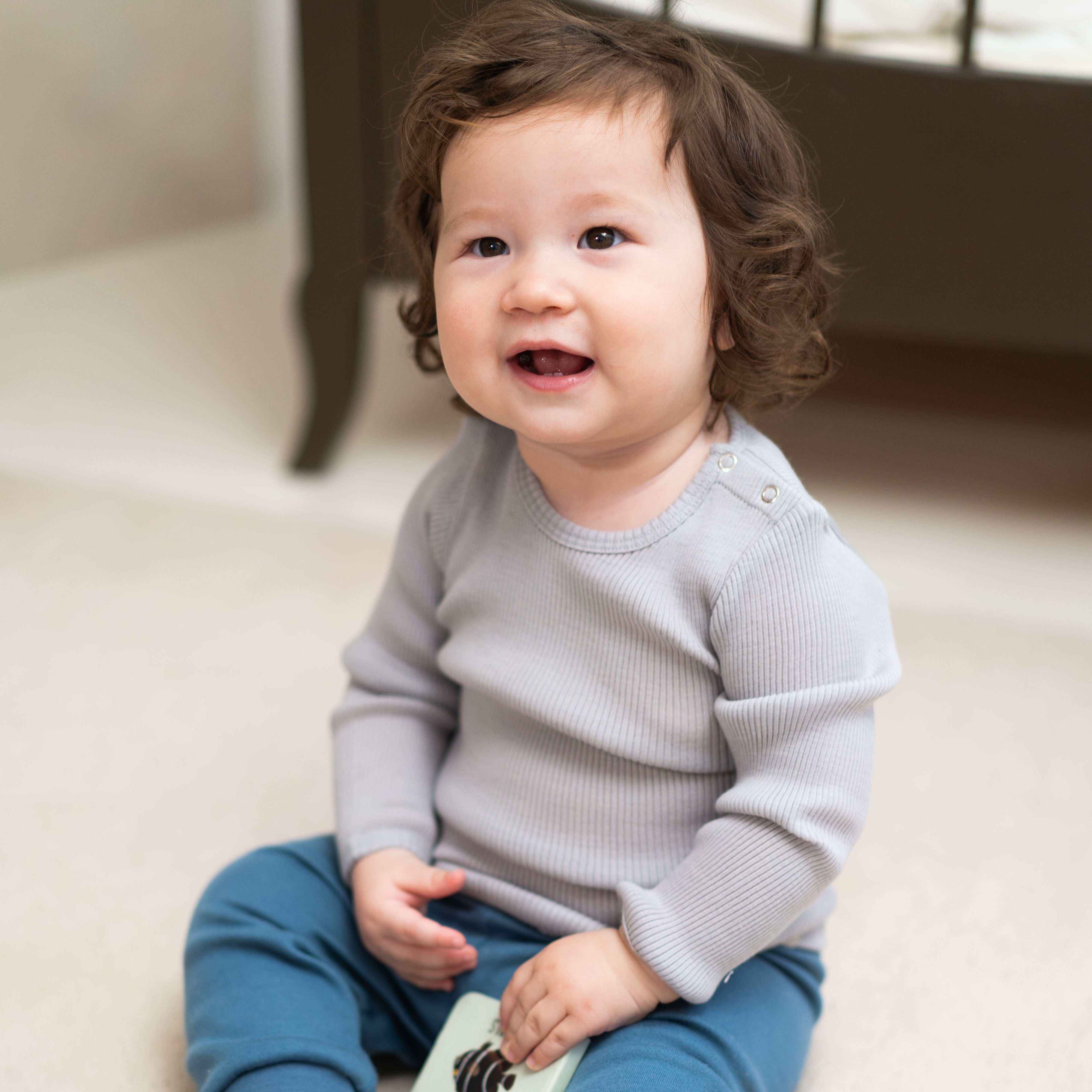 Ein Baby mit lockigem braunem Haar sitzt auf dem Boden und lächelt ganz entspannt. Es trägt ein graues Langarmshirt und blaue Minimalisma-Leggings aus Bio-Baumwolle und wirkt vor dem Hintergrund eines dunkelbraunen Möbelstücks glücklich und zufrieden.