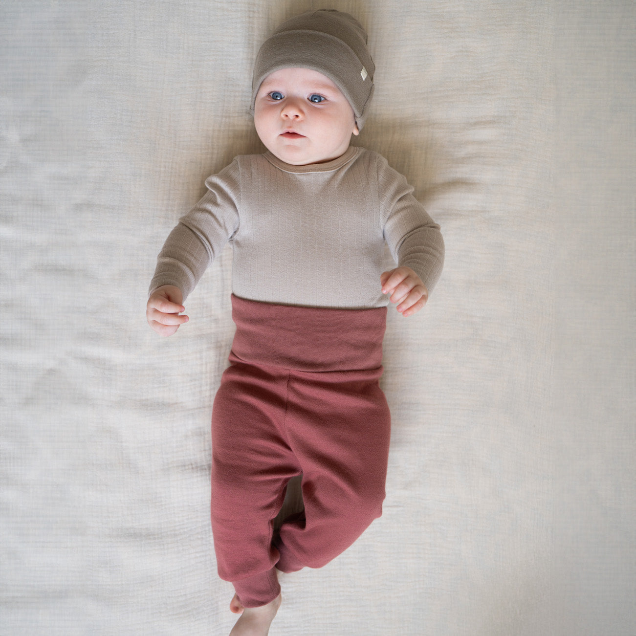 Ein Baby liegt auf einer weichen Unterlage und trägt ein graues Langarmshirt aus Bio-Baumwolle, eine graue Beanie und bordeauxrote Leggings von Minimalisma. Das Baby blickt mit leicht ausgestreckten Armen nach oben und genießt den angenehmen Tragekomfort.