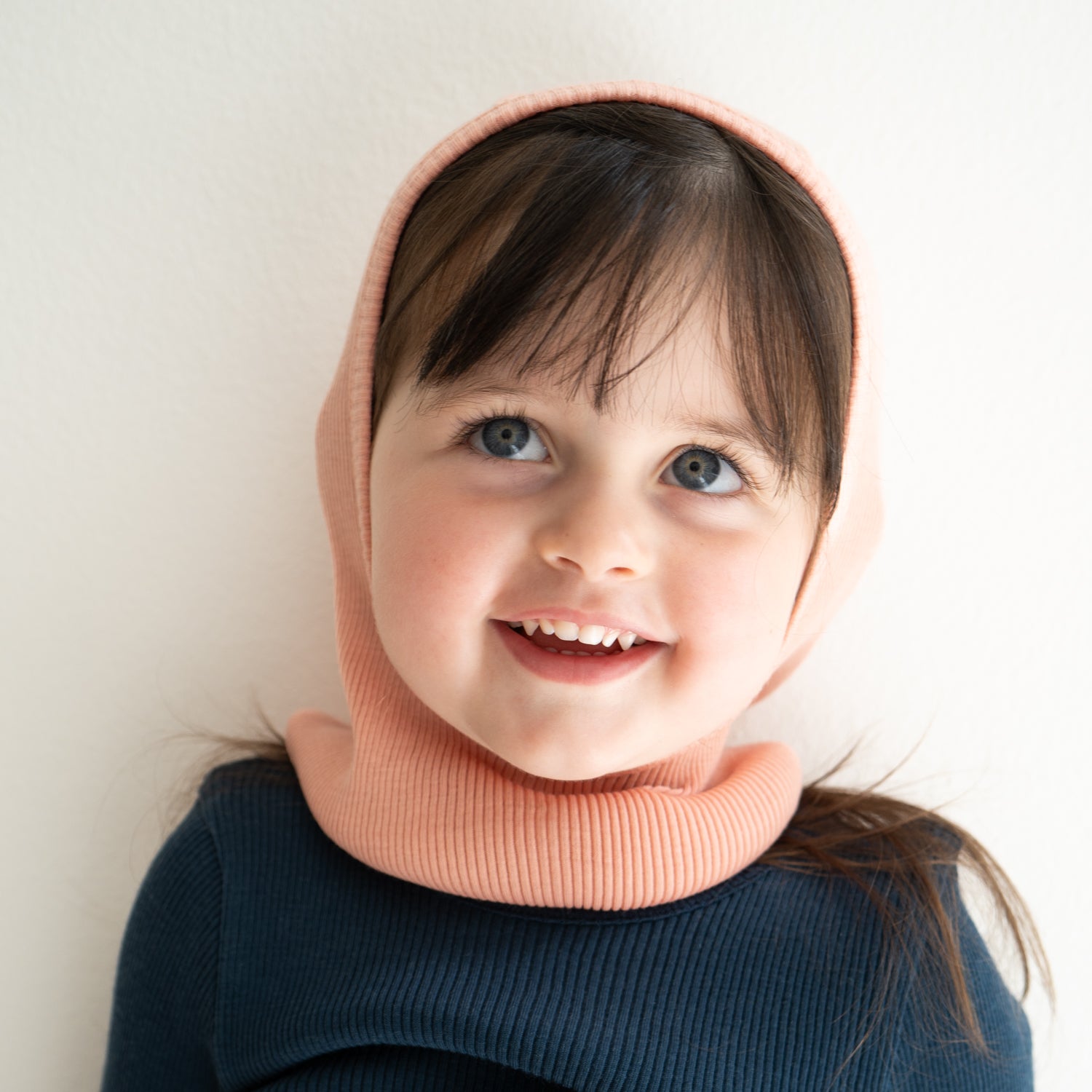 Ein lächelndes kleines Kind mit blauen Augen und dunklem Haar, das vor einem schlichten weißen Hintergrund eine pfirsichfarbene, gerippte Sturmhaube von Minimalisma – Mütze aus antibakterieller Merinowolle und ein marineblaues Hemd trägt.