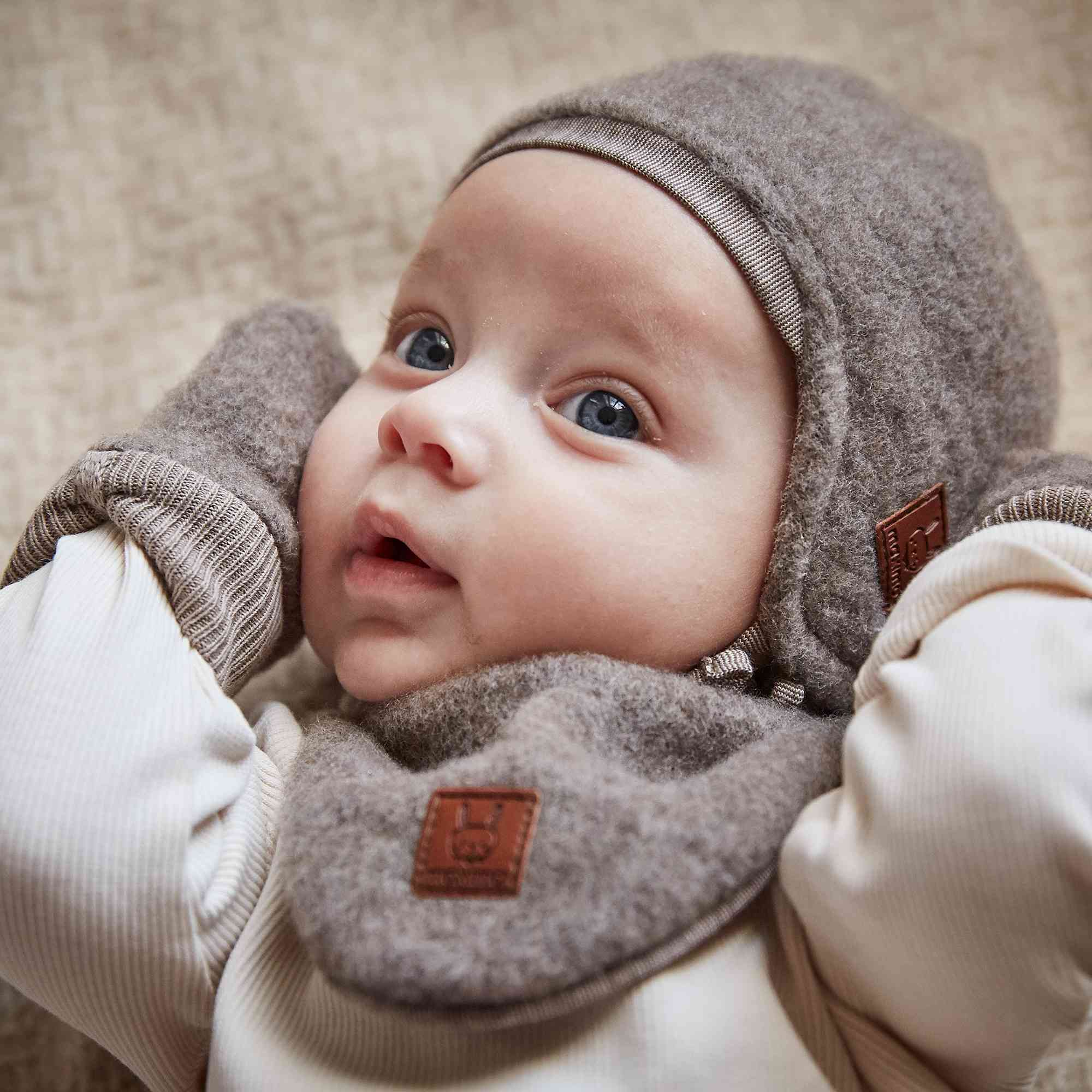 Ein Baby mit blauen Augen liegt da und trägt eine graue Strickmütze, dazu passende Maximo - Babyhandschuhe aus Wollfleece-Fäustlinge und ein strukturiertes Lätzchen mit einem kleinen Lederanhänger. Das Baby schaut nach oben und hat die Hände in der Nähe des Gesichts. Der Hintergrund hat eine beige, gewebte Textur.