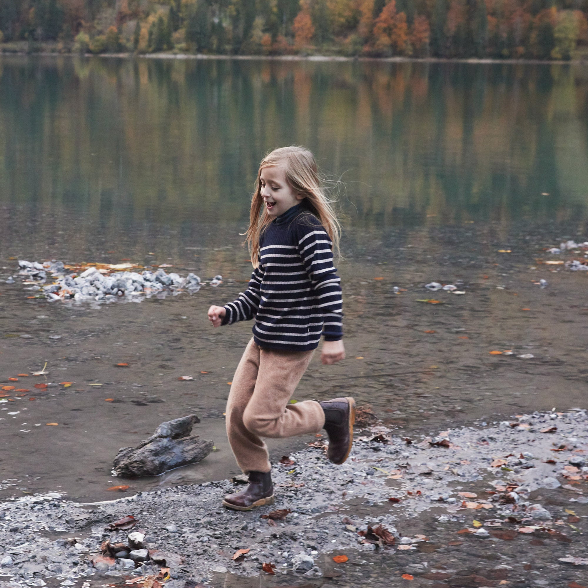 Ein junges Mädchen mit langen Haaren, das ein minimalistisches Langarmshirt und eine hellbraune Hose trägt, läuft fröhlich am felsigen Ufer eines Sees entlang. Im Hintergrund sind ruhiges Wasser, in dem sich Herbstlaub und ein bewaldeter Hügel spiegeln, zu sehen. Ihre gestreifte Bluse aus Merinowolle, zertifiziert nach dem Responsible Wool Standard, hält sie angenehm warm.