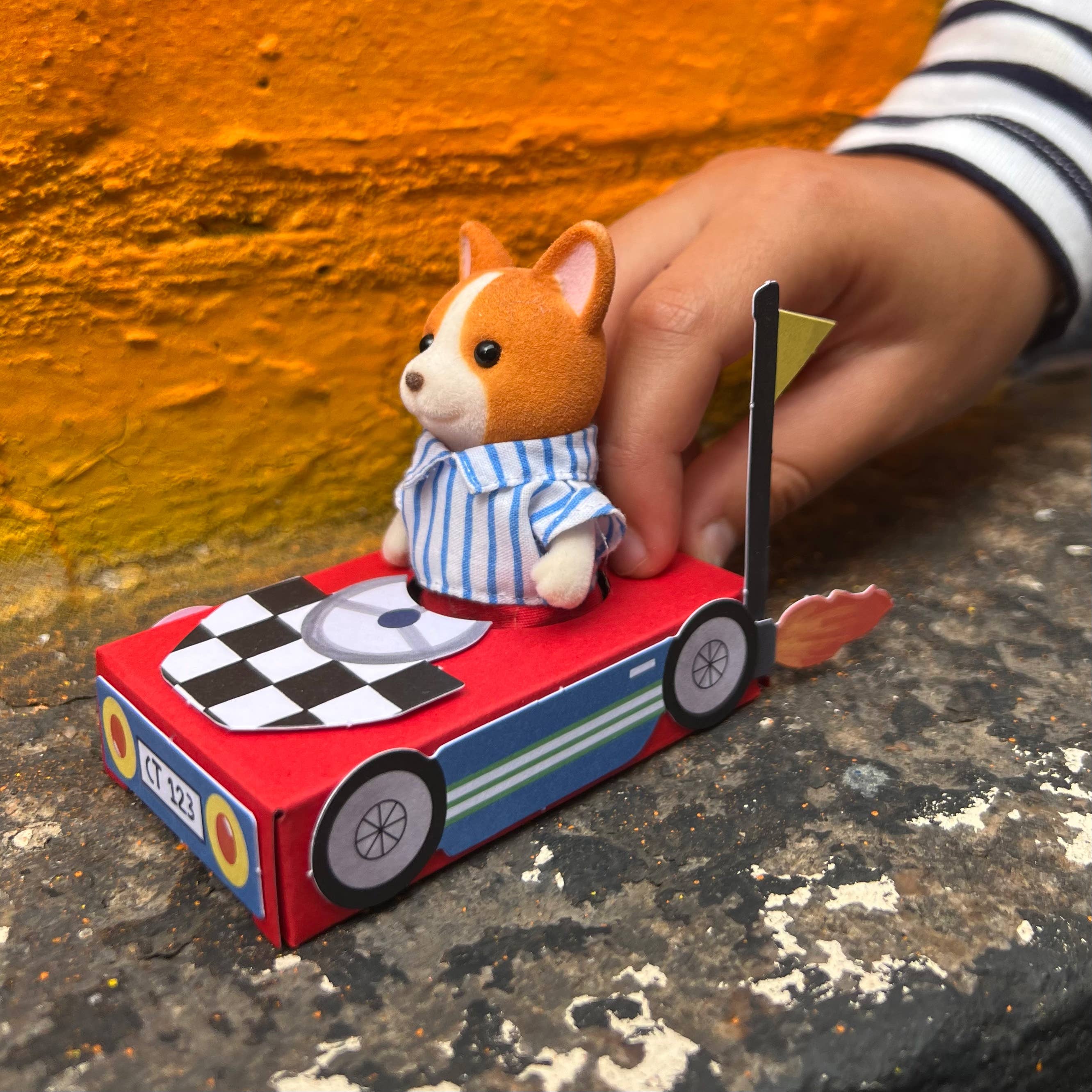 Eine Kinderhand schiebt den Cotton Twist - DIY Matchbox-Rennwagen aus nachhaltigen Materialien, auf dem ein kleiner Spielzeughund in einem blau gestreiften Hemd sitzt. Die Karosserie des Wagens ist rot mit einem karierten Muster auf der Motorhaube und einer gelben Flagge. Der Hintergrund zeigt eine orangefarbene, strukturierte Wand.