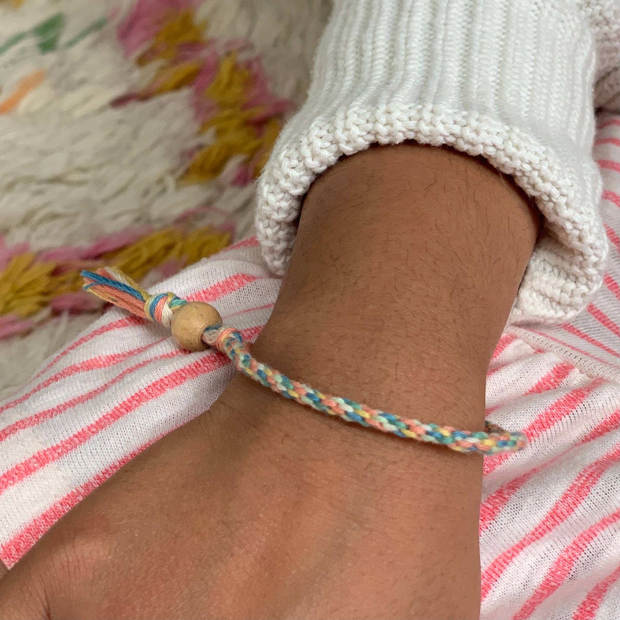 Eine Nahaufnahme des Handgelenks einer Person, die ein handgefertigtes geflochtenes Armband aus dem Cotton Twist - DIY Freundschaftsarmband-Set trägt, mit bunten Fäden und einer Holzperle. Die Person trägt einen weißen Strickpullover und rosa gestreifte Hosen, im Hintergrund ist bunter strukturierter Stoff zu sehen.