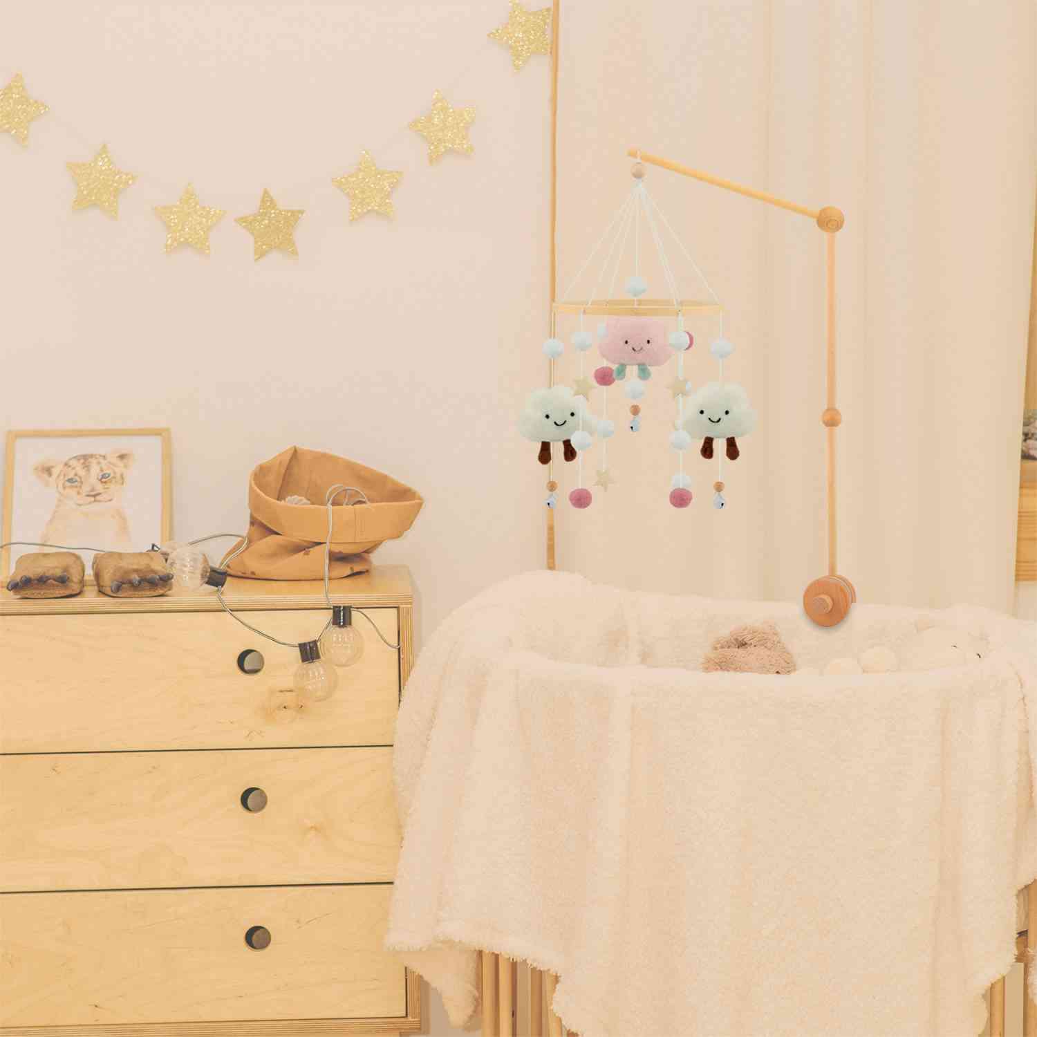 Ein gemütliches Kinderzimmer verfügt über ein Holzbettchen mit einer cremefarbenen Decke und einem Ulysse Couleurs d'Enfance - Mobile mit rosa Wolken von Ulysse Couleurs d'Enfance. Über dem Bettchen zieren goldene Glitzersterne die Wand. Daneben befindet sich eine Holzkommode mit Dekoartikeln, darunter ein gerahmtes Bild eines Löwenjungen und einige ökologische Spielzeuge.