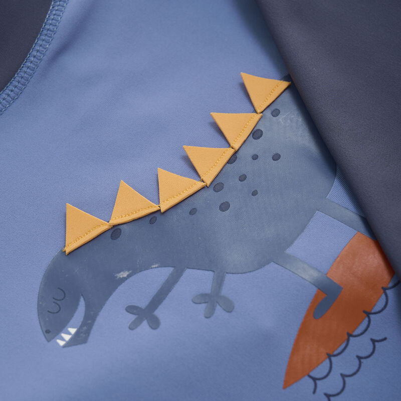 Eine Nahaufnahme eines blauen Stoffes mit einem verspielten Design eines dunkelgrauen Dinosauriers mit orangefarbenen dreieckigen Stacheln auf dem Rücken, der auf einem orangefarbenen Surfbrett läuft. Der Dinosaurier hat ein lächelndes Gesicht und geschlossene Augen, während kleine Punkte und Linien seinem Körper Details verleihen – perfekt für Color Kids – UV-Shirt oder UV-Schutz-Kleidung von Color Kids.