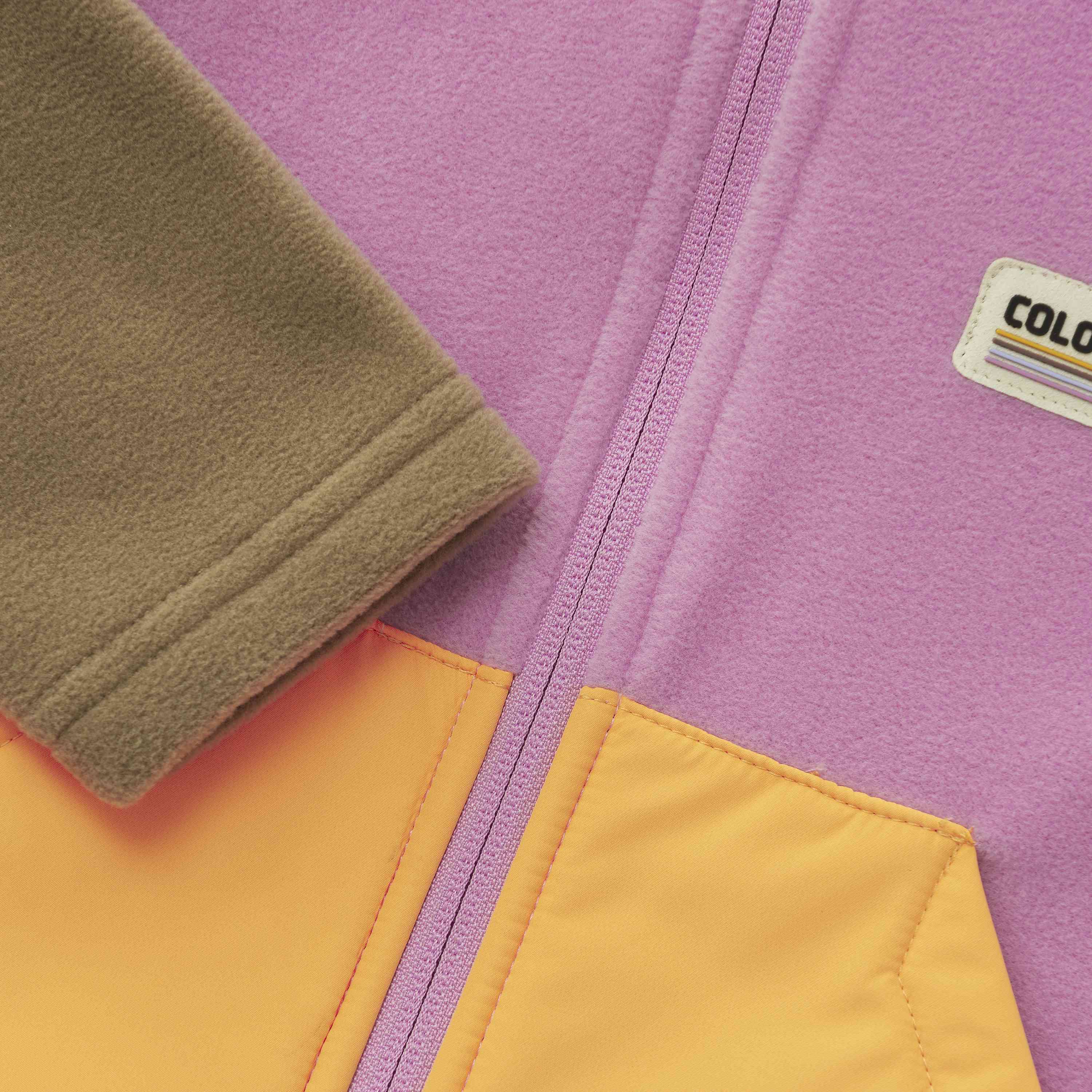 Nahaufnahme einer farbenfrohen Jacke. Die Color Kids - Baby Fleece Jacke von Color Kids besteht aus weichem Fleecematerial mit violettem Körper, gelben Taschen und grünen Ärmeln. In der Mitte verläuft vertikal ein Reissverschluss. Auf der rechten Seite ist ein Etikett mit dem Text „COLORADO“ zu sehen.