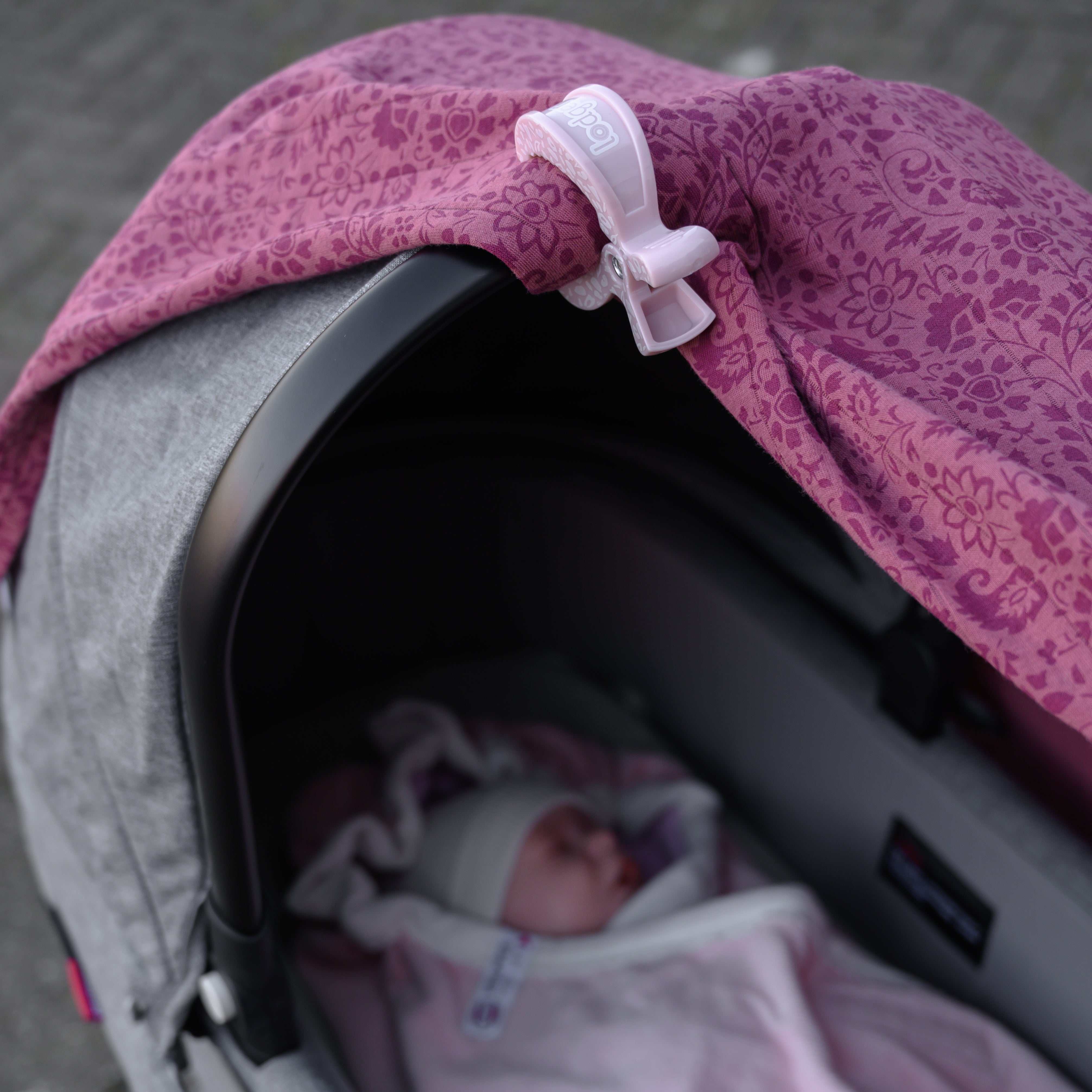 Ein Baby, bekleidet mit einer GOTS-zertifizierten weißen Mütze und einer rosa Decke, schläft friedlich in einem Kinderwagen. Der Kinderwagen verfügt über ein grau-rosa Verdeck mit UV-Schutz, das mit einem Lodger-Swaddler-Clip befestigt ist, der mit Blumenmustern auf dem rosa Stoff verziert ist. Der Hintergrund zeigt eine verschwommene gepflasterte Oberfläche.