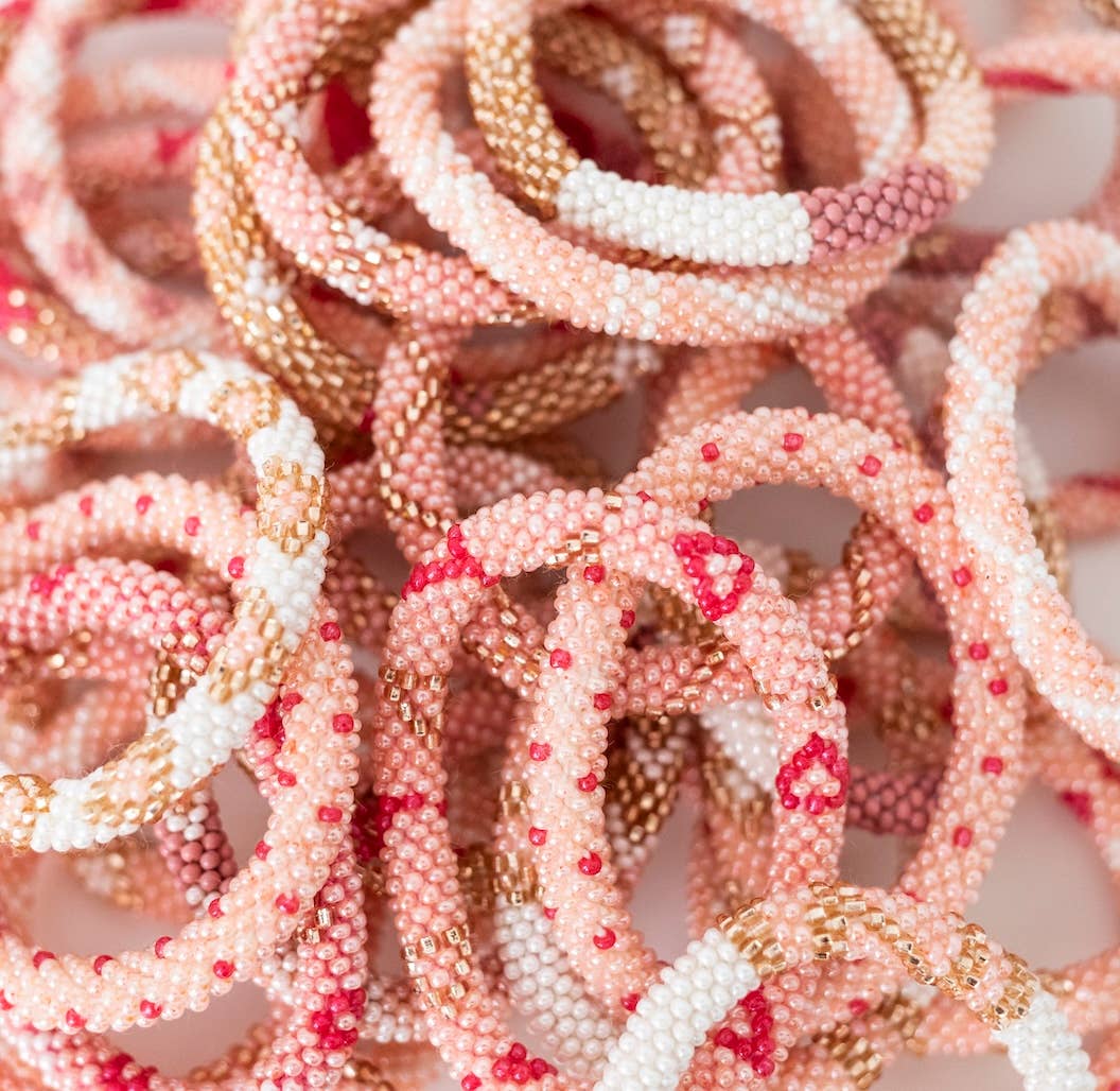 Ein Stapel Aid Through Trade - Armbänder Kinder in verschiedenen Rosa-, Weiß- und Goldtönen. Die Armbänder sind aufwendig mit kleinen Perlen verwoben, wodurch ein strukturiertes und farbenfrohes Muster entsteht. Einige haben Streifen, während andere zufällige Farbspritzer aufweisen, was ihnen ein einzigartiges Aussehen verleiht und die Fair Trade Federation unterstützt.