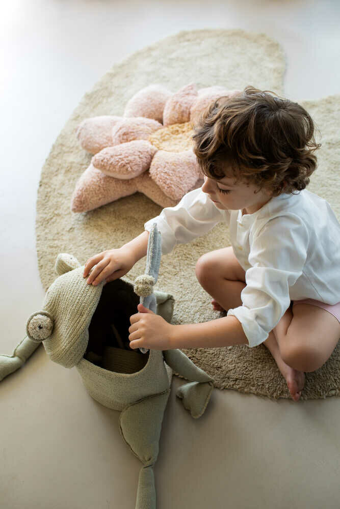 Ein kleines Kind mit lockigem Haar, das ein weißes Hemd und rosa Shorts trägt, sitzt auf einem beigen Teppich in seinem Kinderzimmer. Es spielt mit einem Aufbewahrungskorb „Fred der Frosch“ von Lorena Canals, der einen offenen Mund hat, und legt einen kleinen Gegenstand hinein. Hinter dem Kind ist ein großes, plüschiges Blumenkissen zu sehen, das Teil der Kinderzimmerdekoration ist.