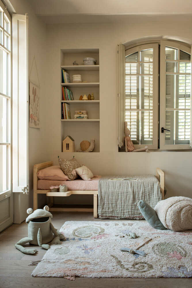 Ein gemütliches Babyzimmer mit einem kleinen Holzbett, rosafarbener Bettwäsche und einer grünen Steppdecke. Das Zimmer verfügt über eingebaute Regale voller Bücher und Spielzeug, Fensterläden und einen flauschigen Teppich mit verspielten Mustern. Als Teil der Einrichtung des jungen Kinderzimmers sind Stofftiere wie Lorena Canals – Aufbewahrungskorb Fred der Frosch von Lorena Canals auf dem Boden verstreut.