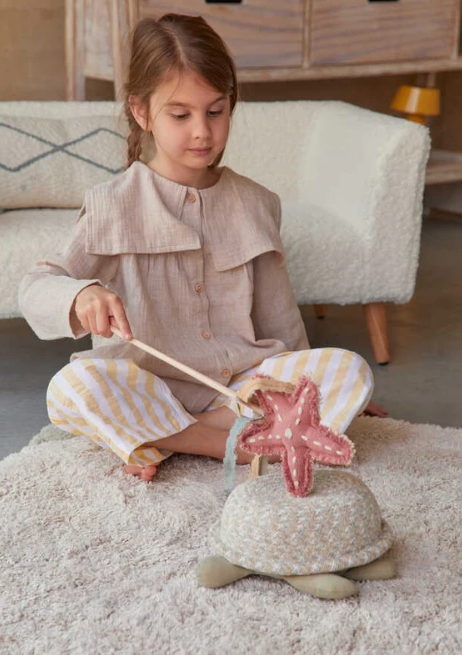 Ein junges Mädchen sitzt auf einem Teppichboden in ihrem Babyzimmer mit Tiermotiven und spielt mit einer Babyschildkröte von Lorena Canals - Aufbewahrungskorb. Sie benutzt einen Holzstab, um mit einem weichen, rosa Seestern zu interagieren, der am Rücken der Schildkröte befestigt ist. Sie trägt ein hellbraunes Oberteil und gestreifte gelb-weiße Hosen.